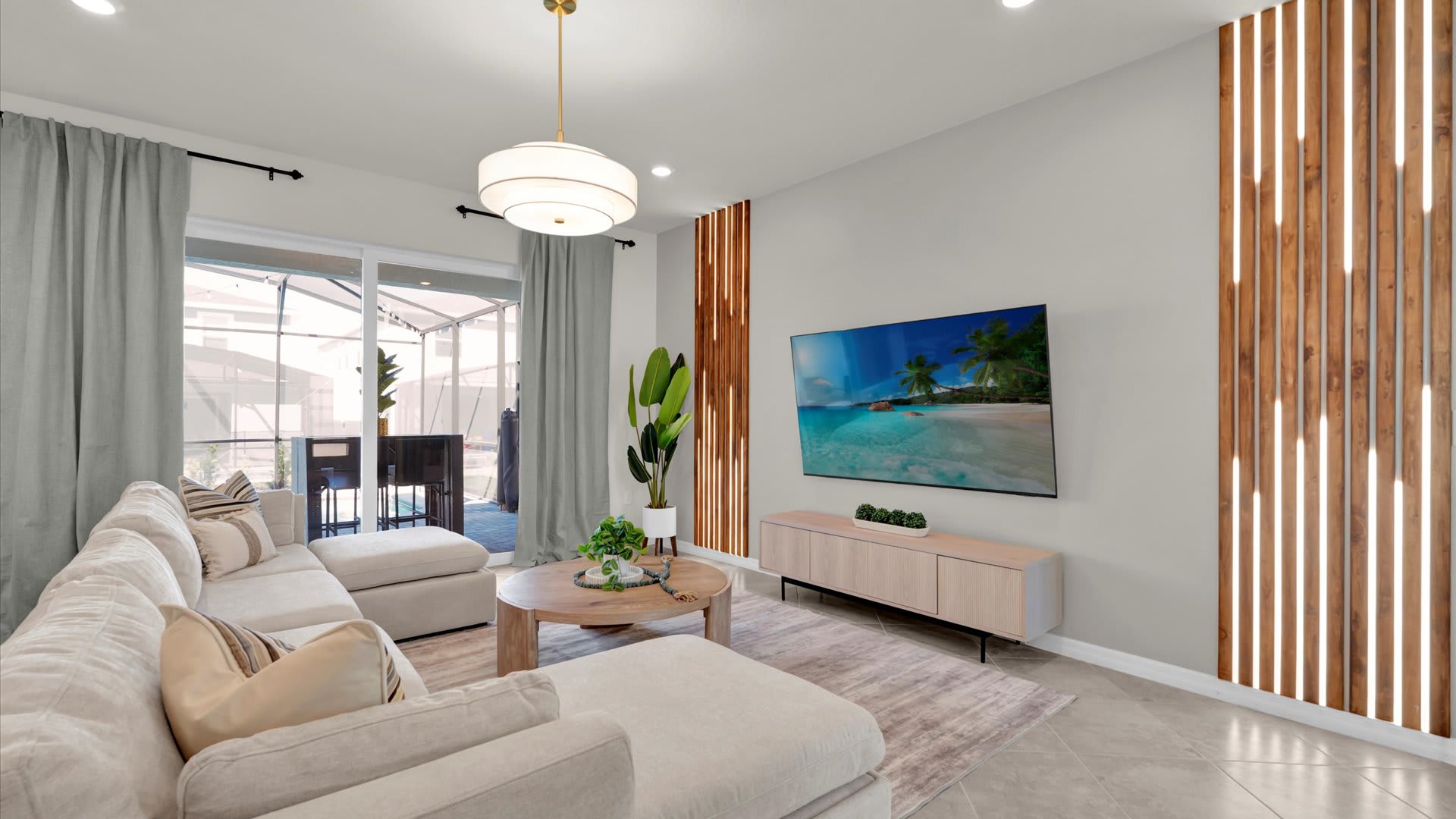 Contemporary living room featuring a large wall-mounted TV above a light wood console, flanked by vertical wooden slat accents. A cream sectional sofa and a round coffee table face sliding glass doors leading to a screened patio.