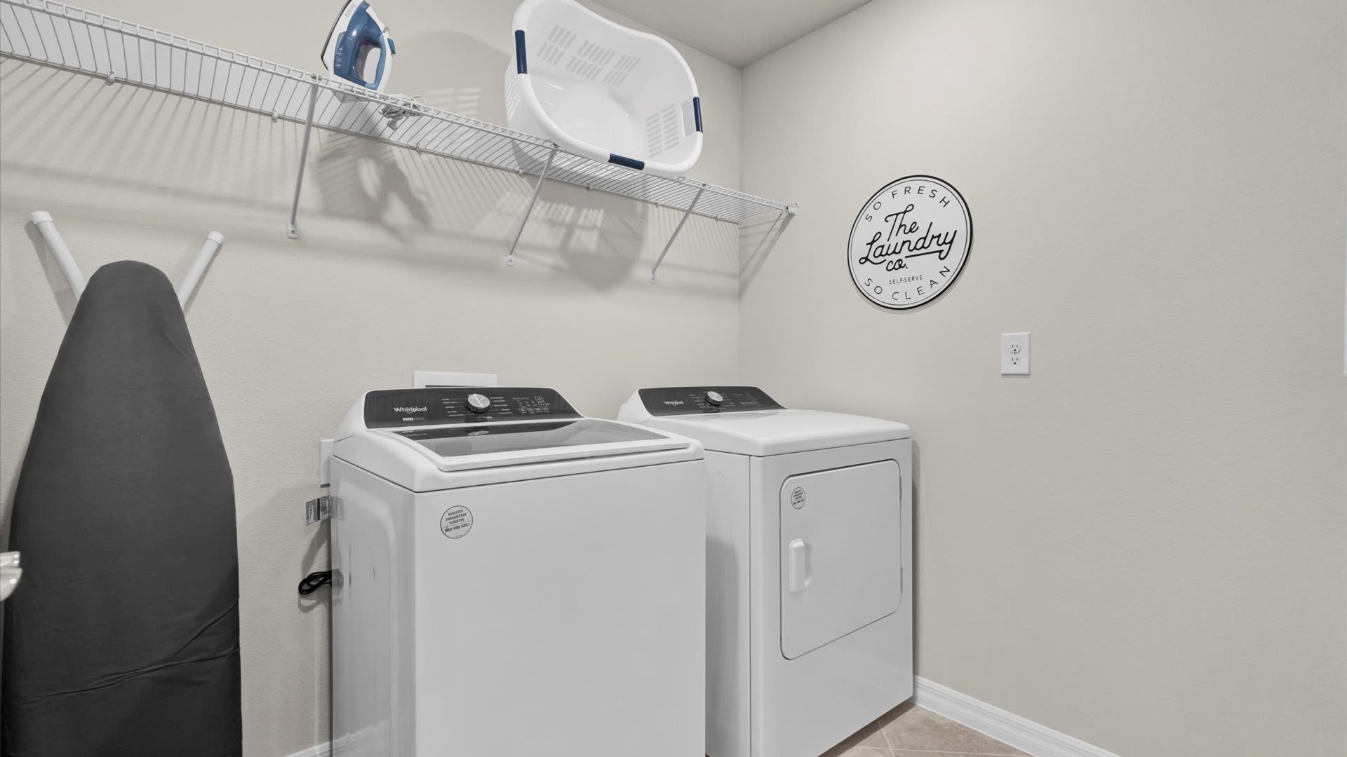 A functional laundry room equipped with a white top-loading washing machine and a front-loading dryer. A wire shelf above holds an iron and a laundry basket, with an ironing board leaning against the wall.