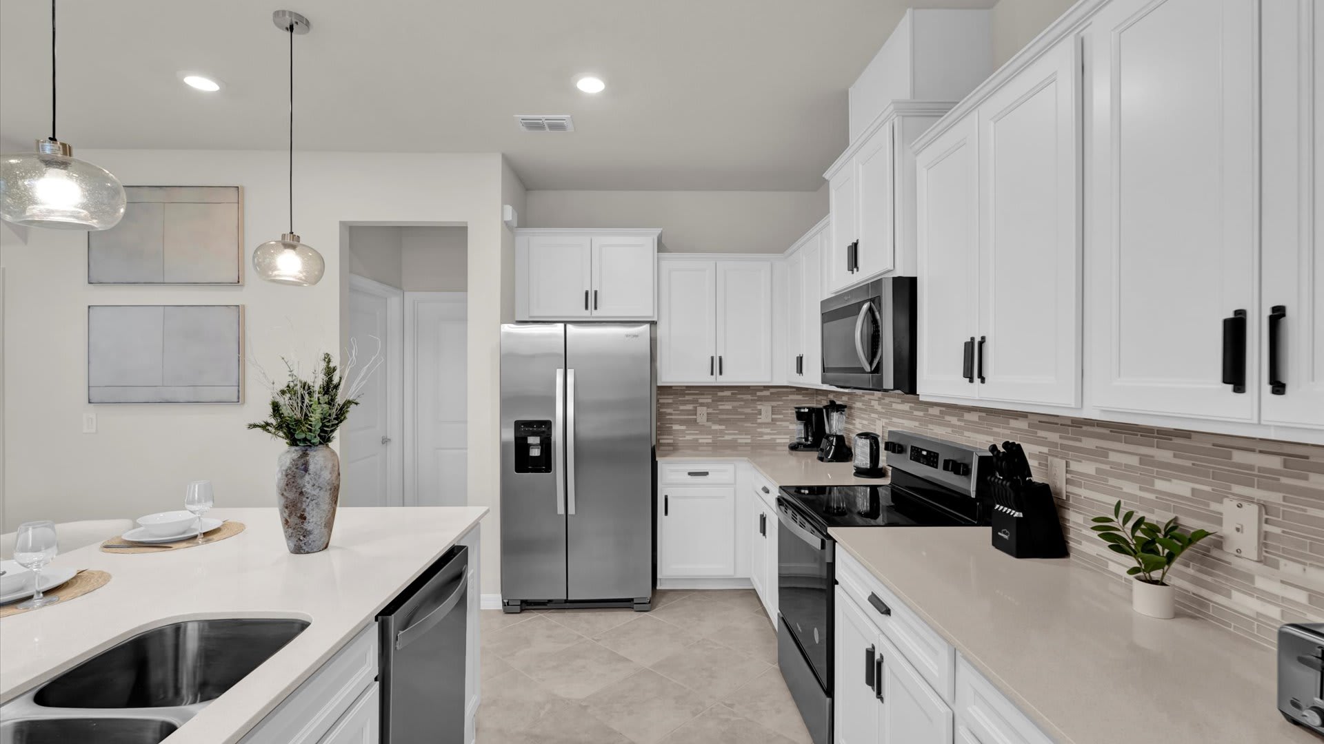 Well-appointed kitchen with white shaker cabinets, light countertops, and a beige mosaic tile backsplash. Stainless steel refrigerator, black range, and microwave are visible, along with small appliances like a coffee maker and toaster.