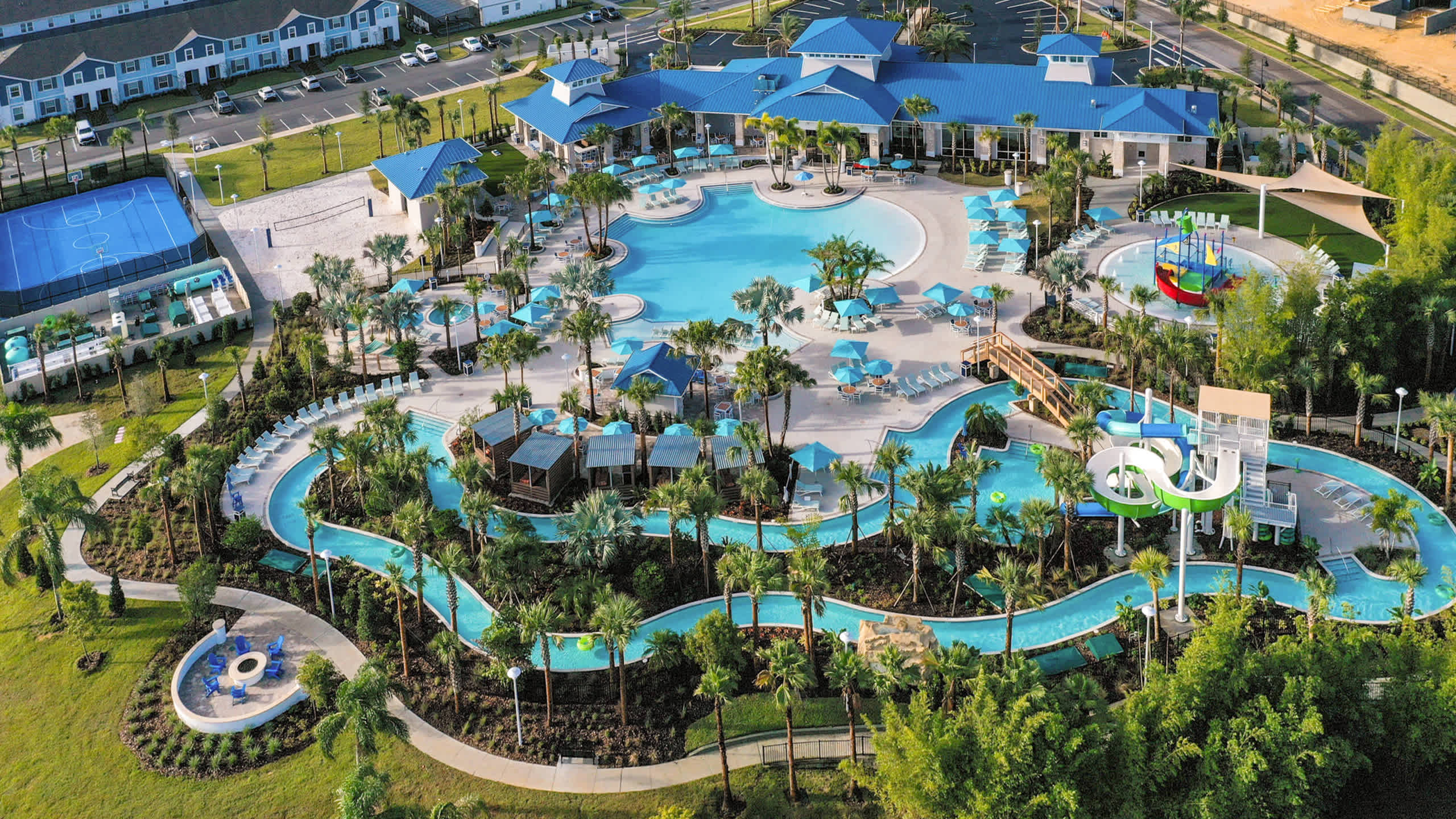 Aerial view of a large resort-style communal pool complex featuring multiple swimming areas, a lazy river, water slides, and numerous lounge chairs with umbrellas. Surrounding the pools are palm trees, green landscaping, and other resort buildings.