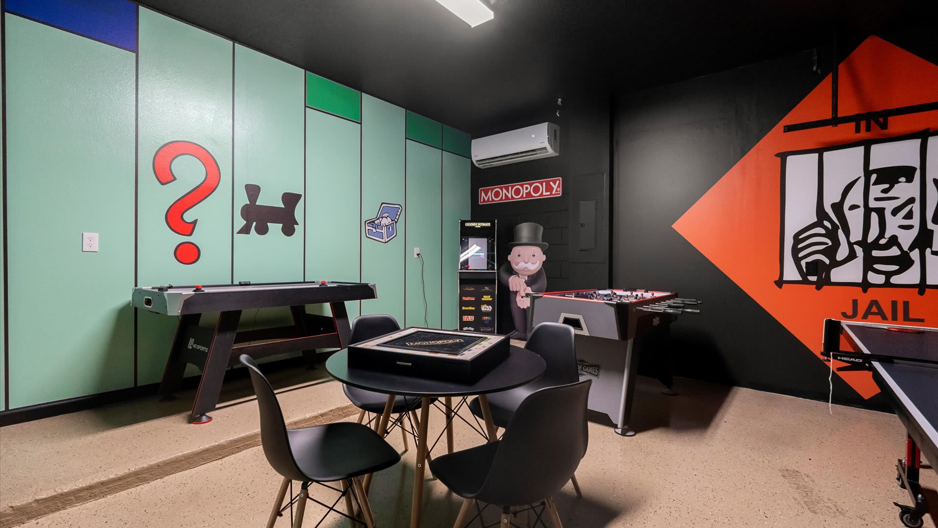 Another perspective of the Monopoly-themed game room, showcasing the light green walls with game board elements and a black ceiling. A ping-pong table is in the foreground, with a round table and chairs, a foosball table, and a pool table in the background.