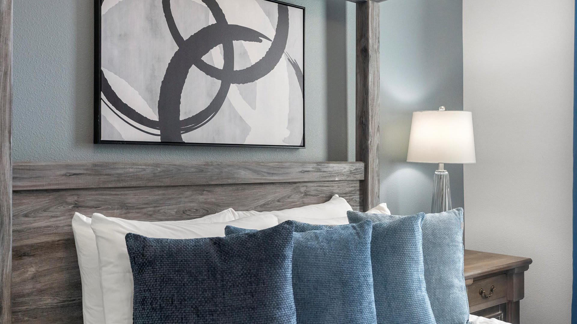 A close-up of a rustic wooden four-poster bed with a gray-washed finish, adorned with various shades of blue decorative pillows, and a modern abstract artwork hanging above the headboard.
