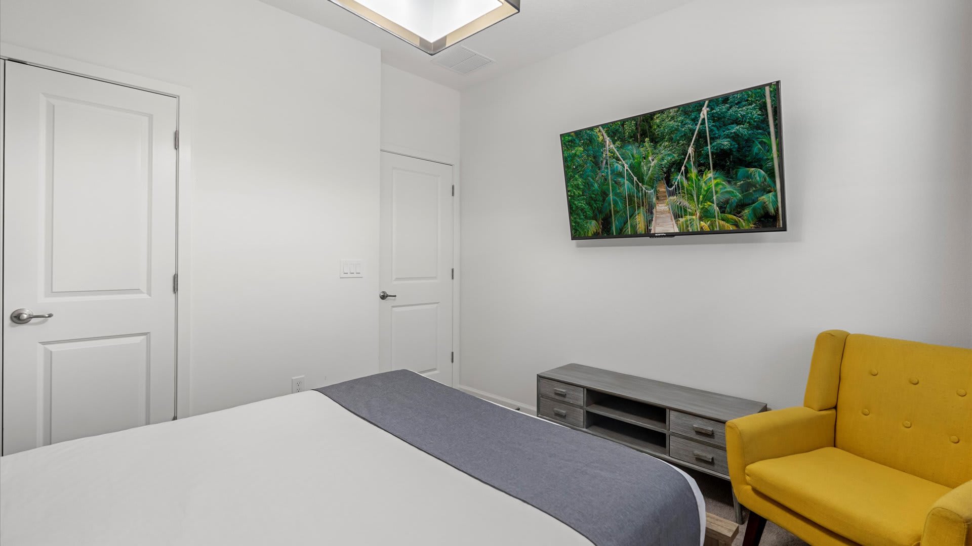 View of bedroom-1 showing a wall-mounted flat-screen TV displaying a jungle scene, a yellow tufted armchair, and a gray dresser. Two white doors are visible on the left.