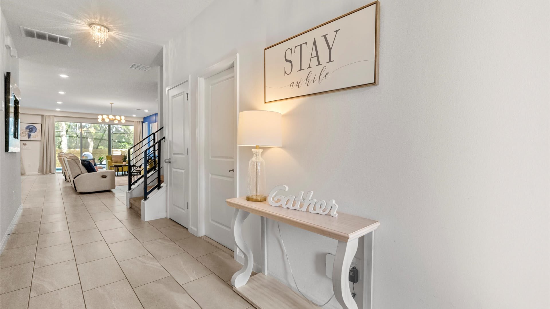 A welcoming foyer with light tile flooring, a console table adorned with a "Gather" sign and a lamp, and a large "Stay a while" sign on the wall, leading to an open living space.