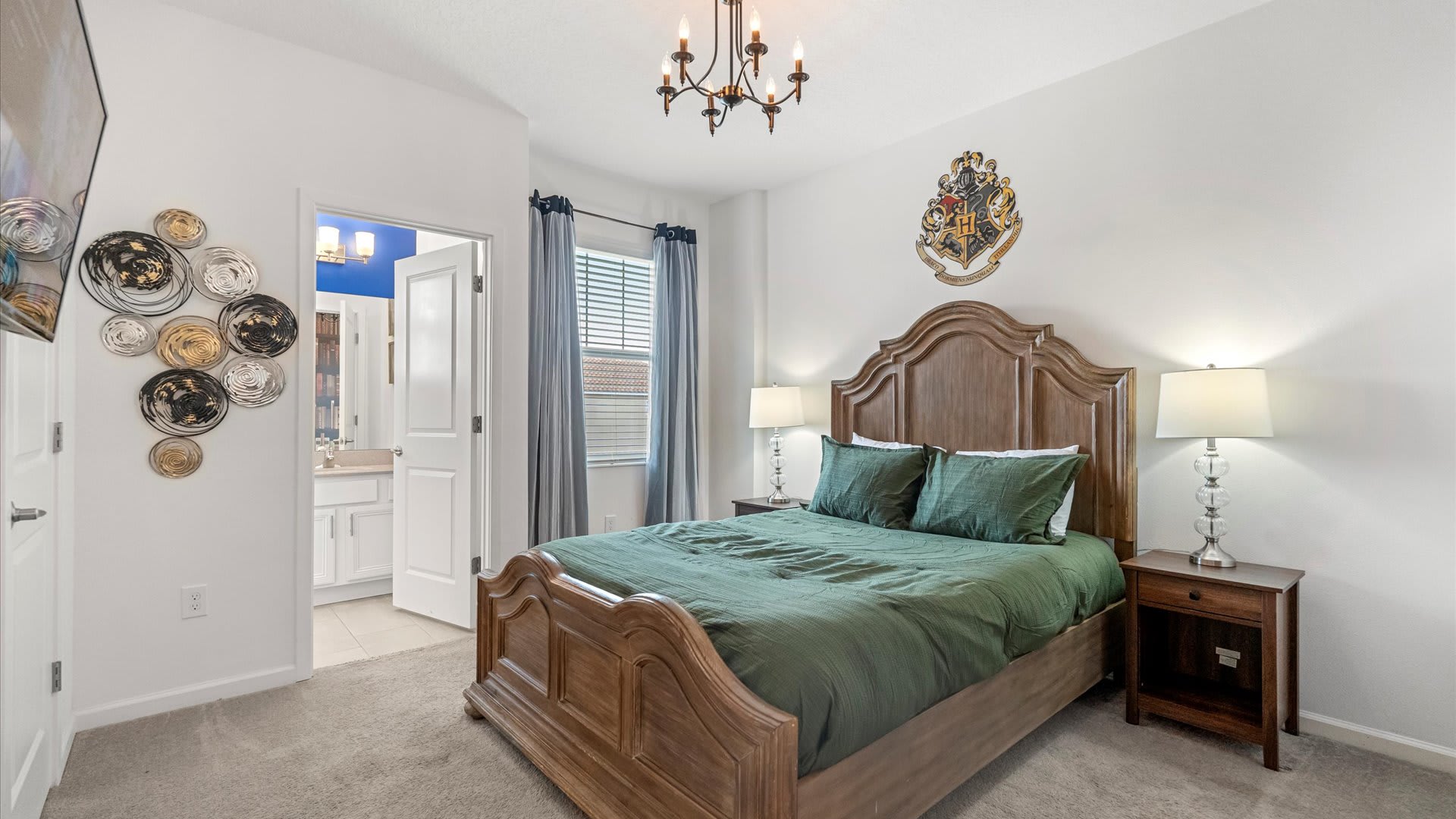 Harry Potter themed bedroom with a dark wood sleigh bed, green bedding, two nightstands with lamps, a chandelier, and a large Hogwarts crest on the wall. An open door leads to a bathroom.