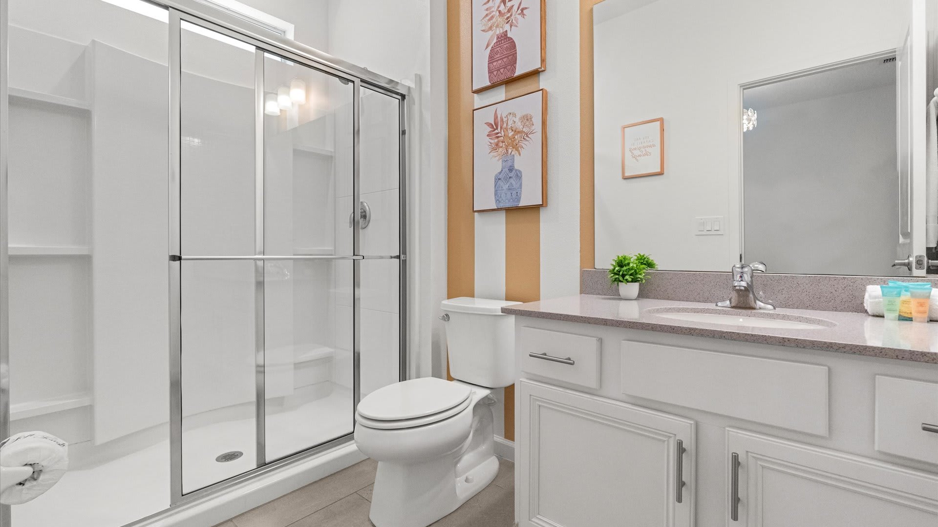 Modern bathroom with a glass-enclosed shower, white toilet, and a single vanity with a light gray countertop. The wall behind the toilet features vertical orange and white stripes.