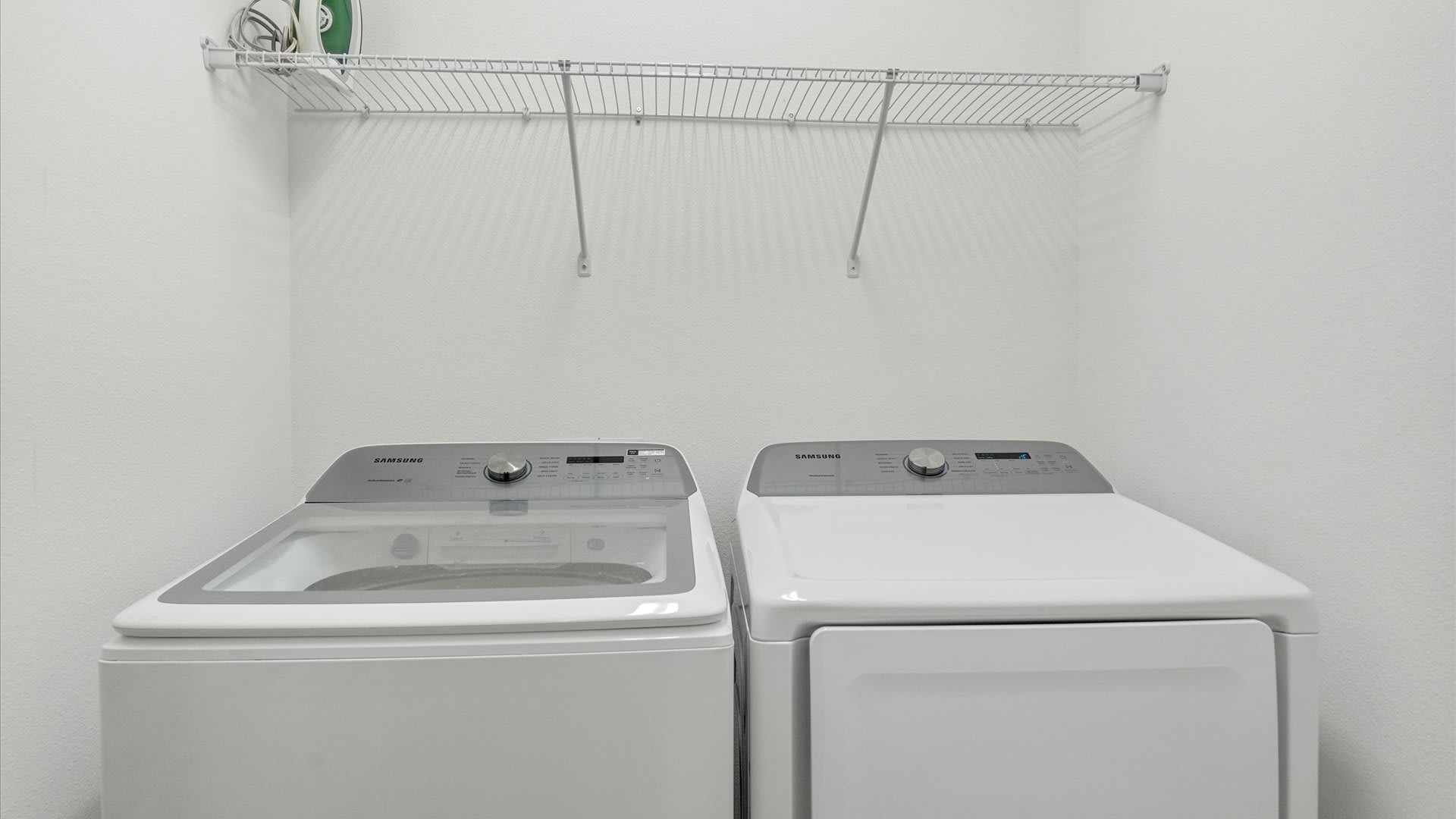 Functional laundry room equipped with a white Samsung top-load washer and front-load dryer, a wire shelf for storage, and an ironing board.