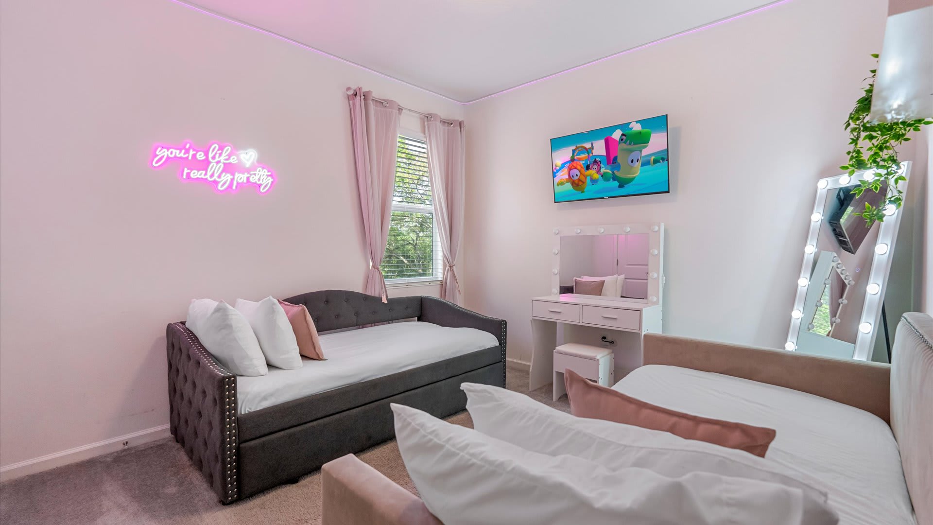 Pink-themed bedroom designed for children or teens, featuring a tufted gray daybed, a second bed with a velvet headboard, a vanity with a lighted mirror, and a neon sign that reads "you're like really pretty."