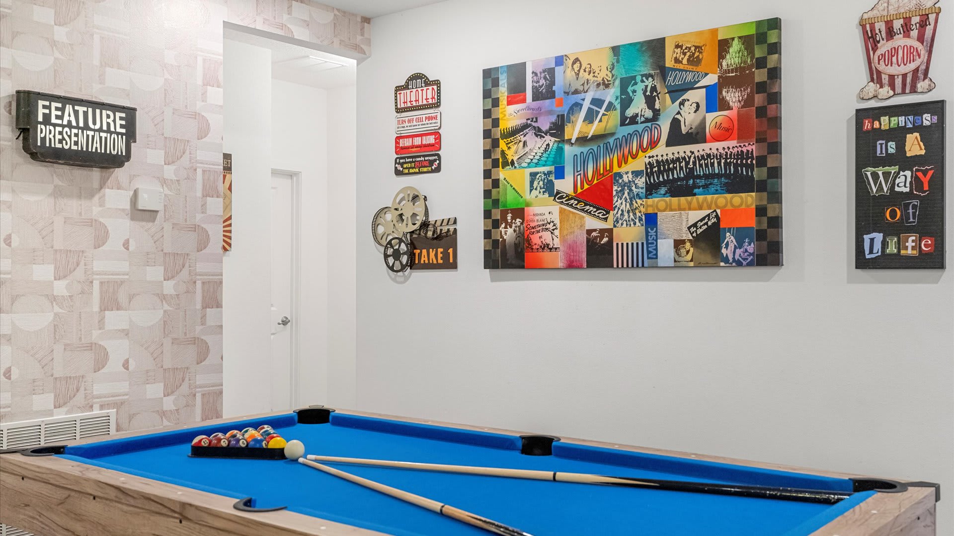A close-up of the game room highlights a light wood pool table with blue felt, cue sticks, and billiard balls, surrounded by movie-themed wall decor including a 'FEATURE PRESENTATION' sign and a vibrant Hollywood collage.