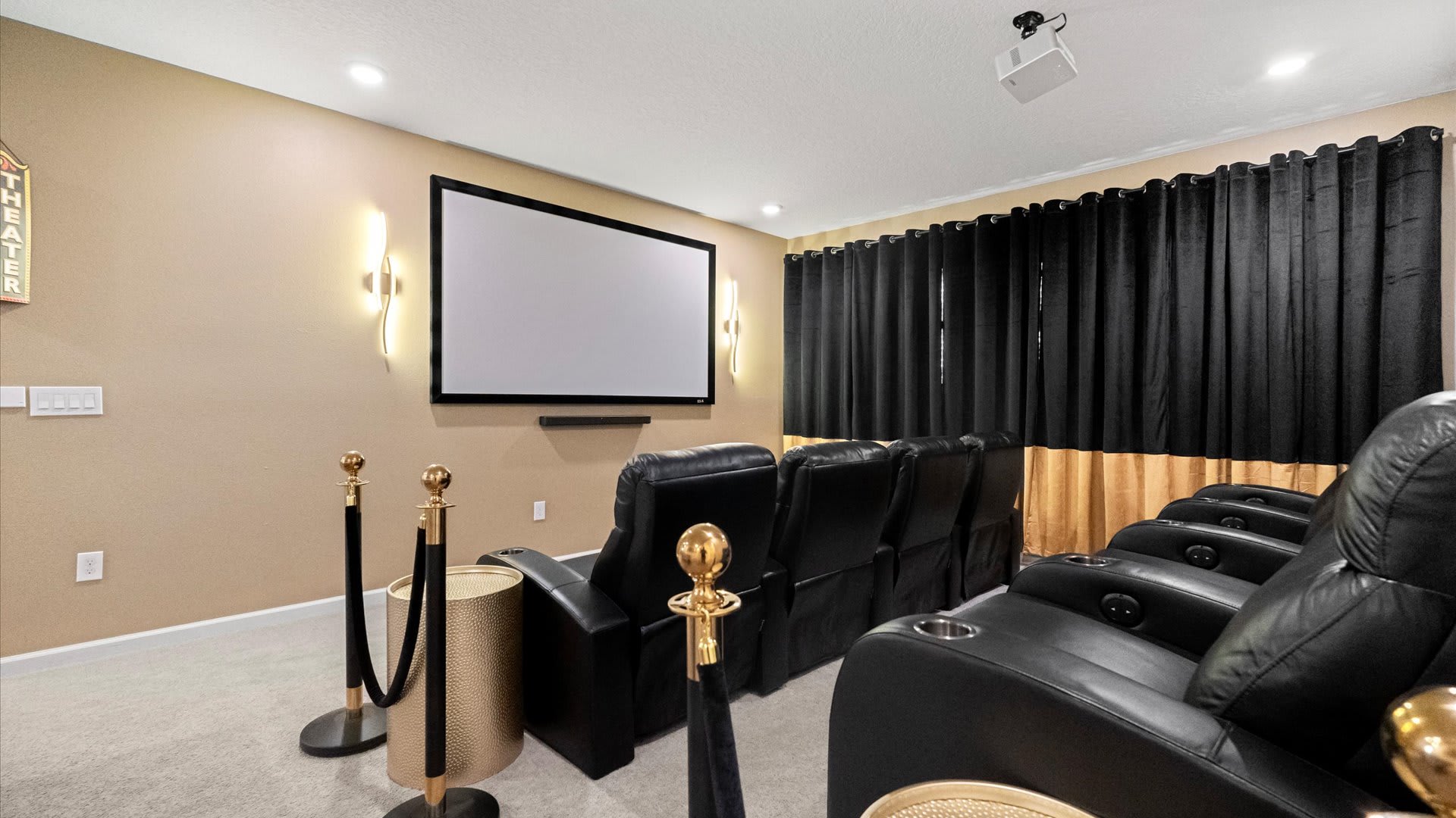 A dedicated home theater features a large projection screen, a ceiling-mounted projector, and multiple rows of plush black reclining chairs, flanked by gold stanchions and black room-darkening curtains.