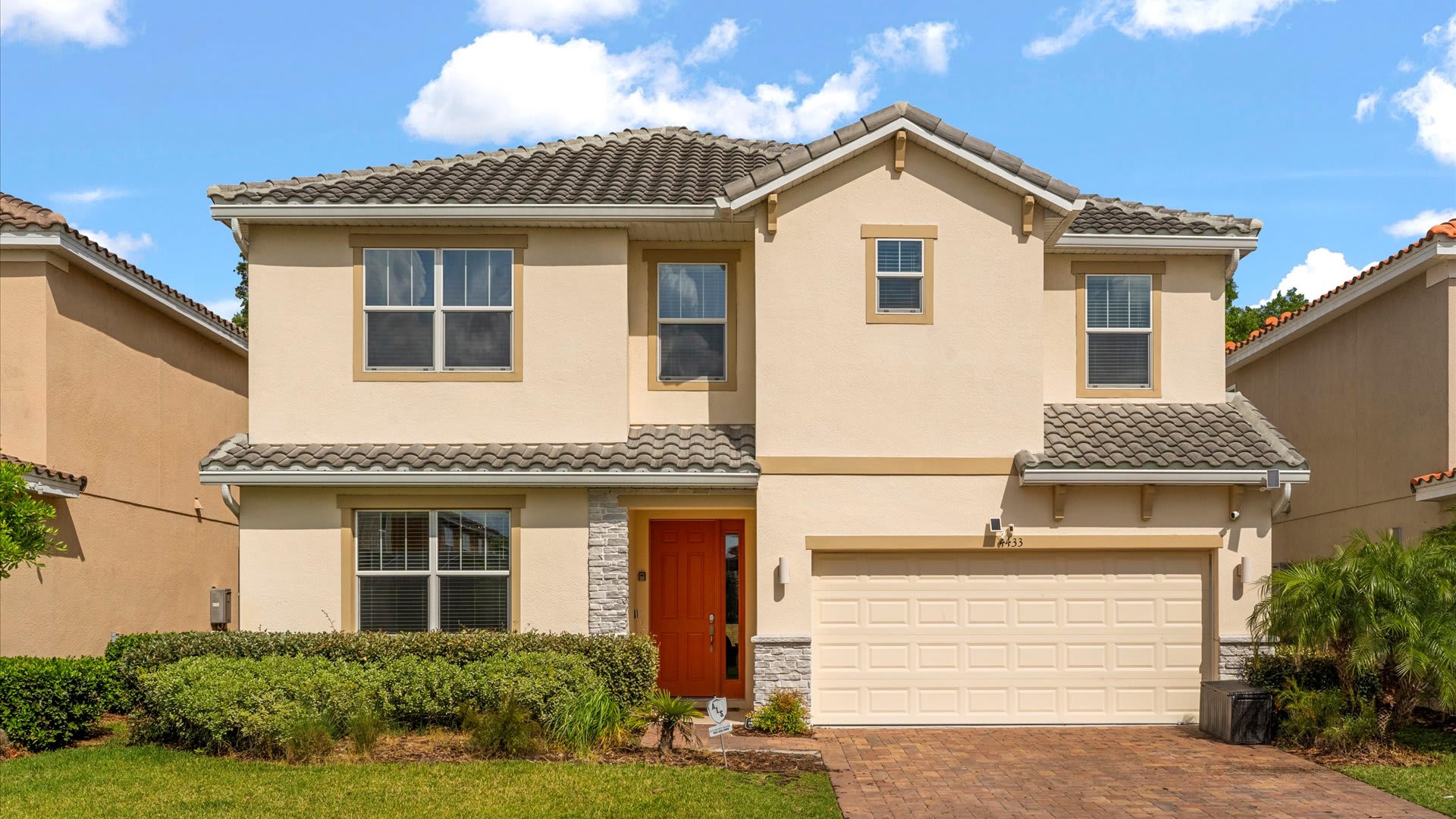 The front exterior of a two-story house with a light beige stucco finish and a tiled roof. It features an orange front door, a two-car garage, and well-maintained landscaping with green bushes.
