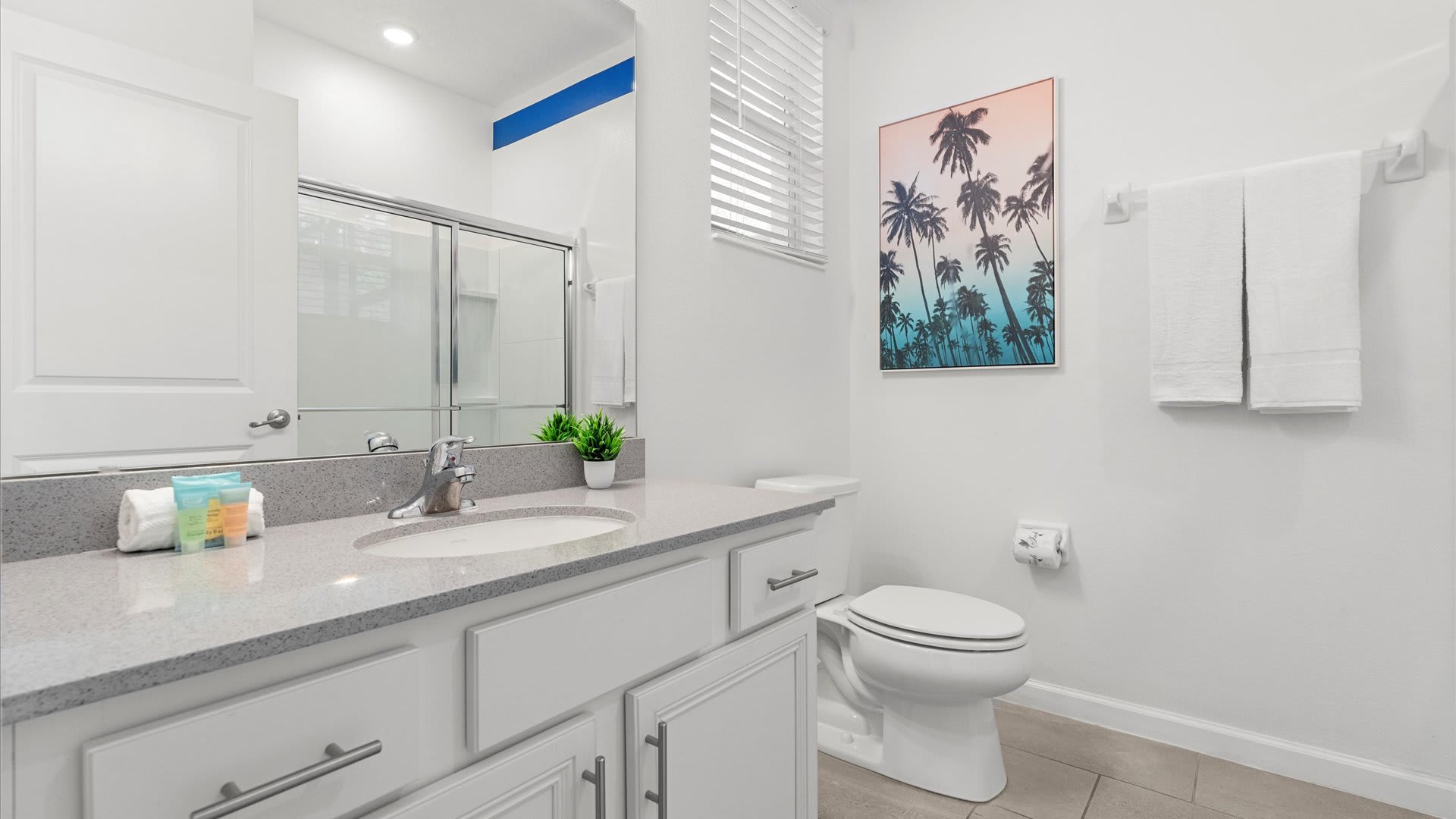 Bright white bathroom with a single vanity featuring a light gray countertop, a large mirror, a toilet, and a glass-enclosed shower/tub combo. A framed palm tree print adds a touch of decor.