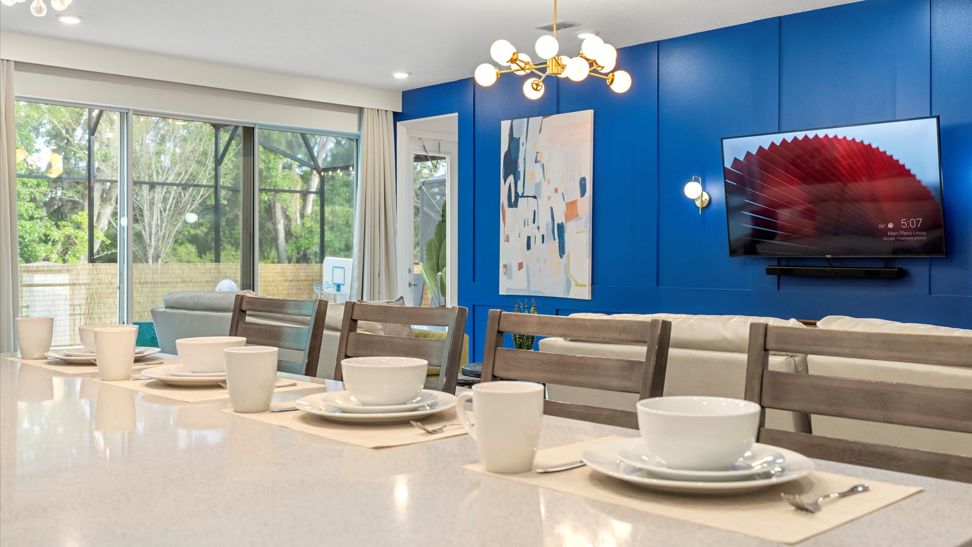 A modern dining or kitchen island area with white place settings, featuring a striking blue accent wall with vertical paneling, a gold Sputnik-style chandelier, and a large TV, overlooking a screened patio.