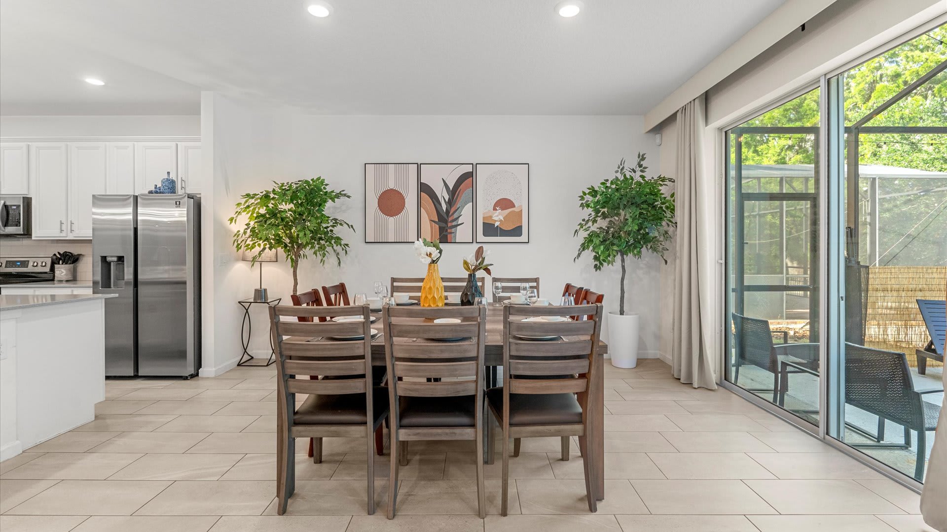 A spacious dining area with a long wooden table and chairs, positioned between two large artificial plants, with a view of the modern kitchen and sliding glass doors to the outdoor space.