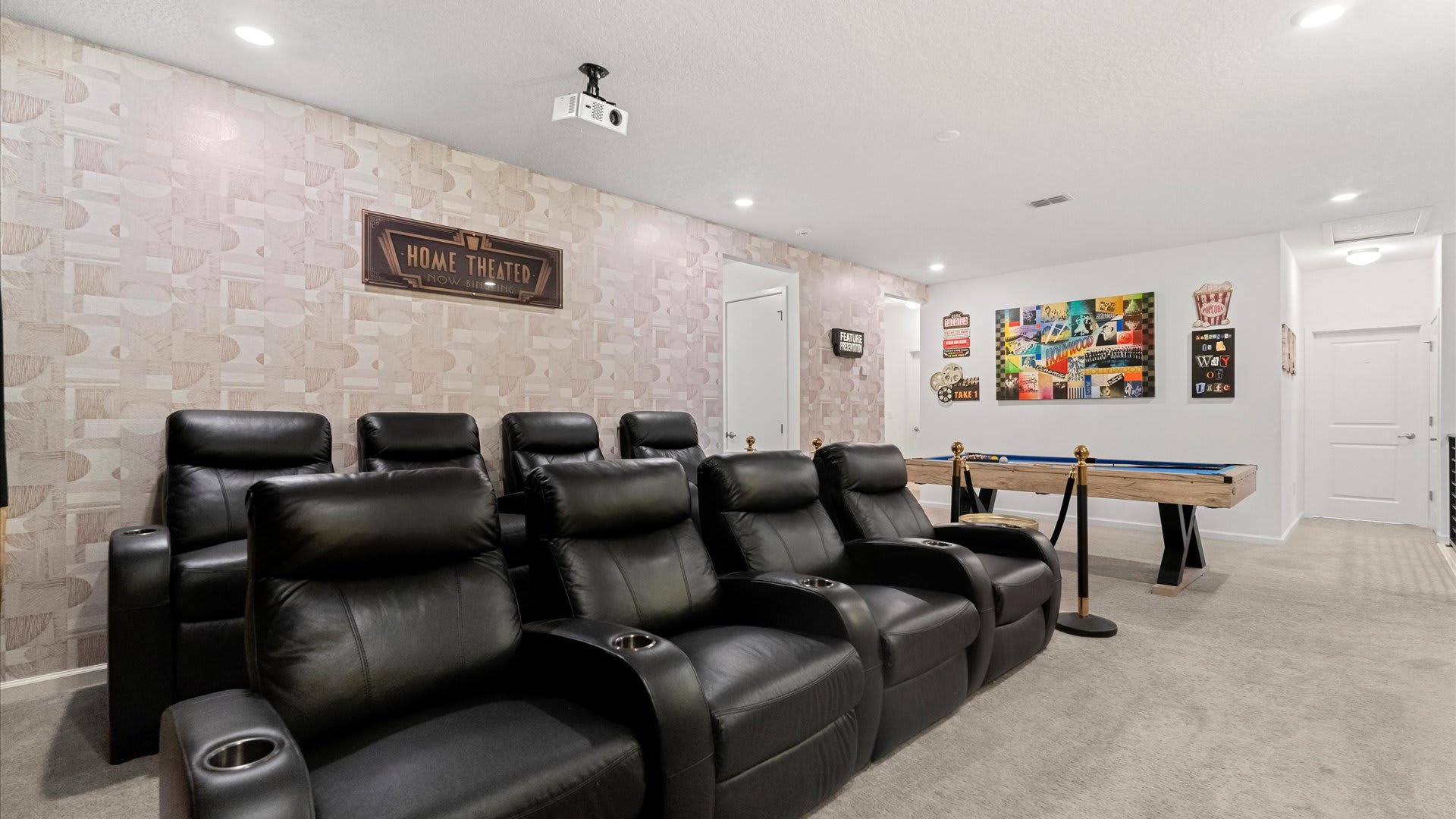 A spacious game room features multiple rows of black leather reclining theater seats facing a movie-themed wall, with a light wood pool table with blue felt in the foreground, set against patterned wallpaper.