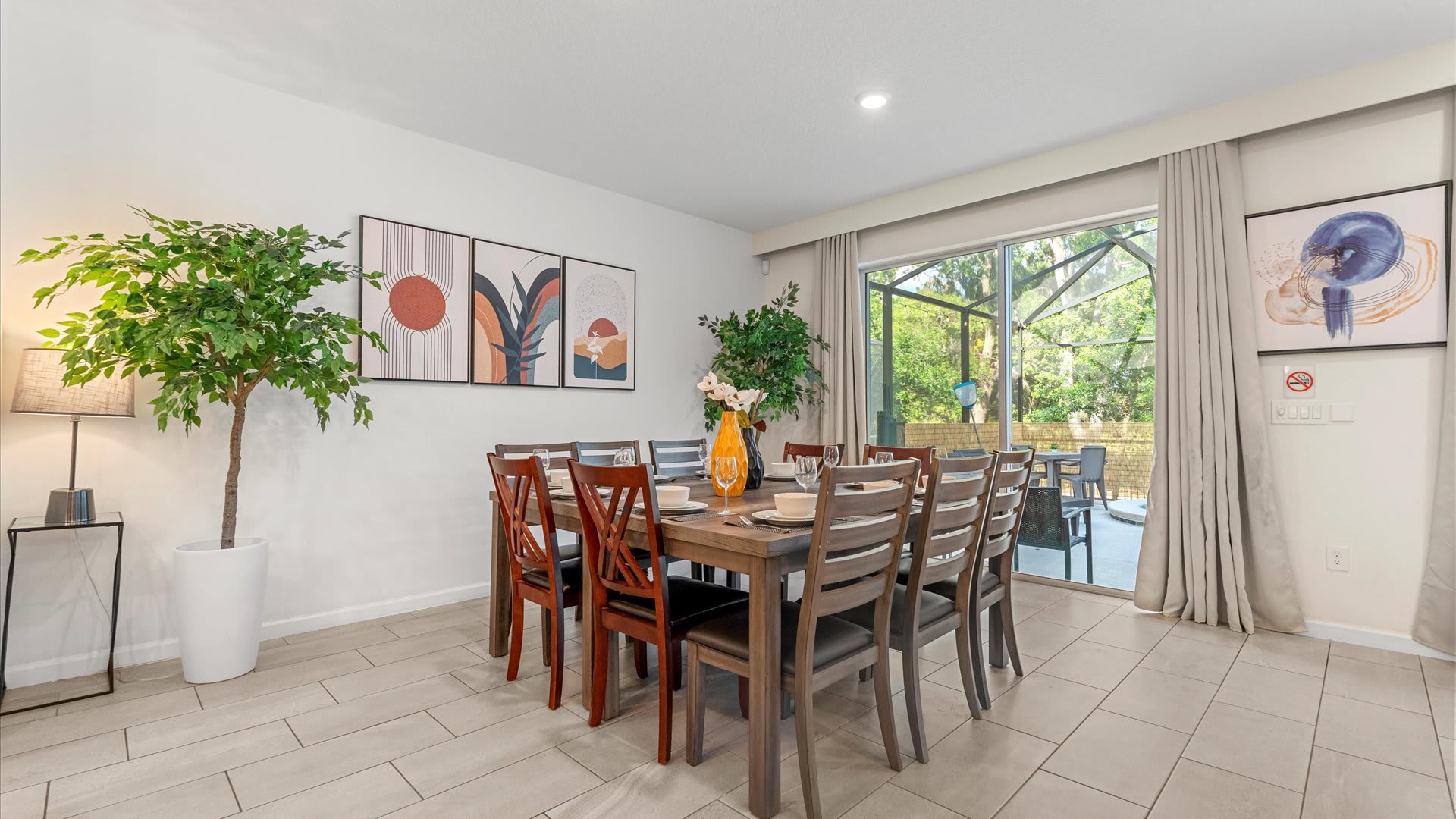 A bright dining room featuring a large wooden table with seating for ten, flanked by two tall artificial trees, with sliding glass doors leading to an outdoor patio area.