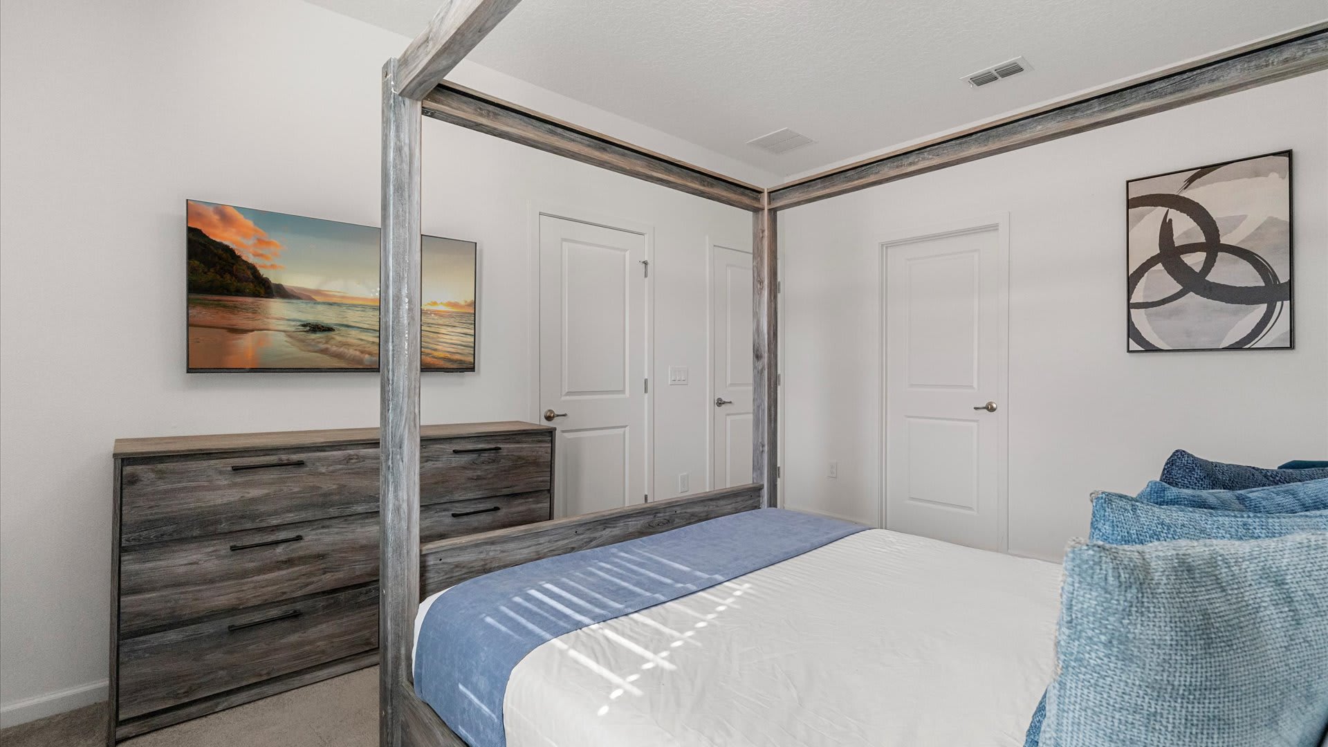 Bright bedroom featuring a rustic wooden four-poster bed with blue and white bedding, a matching dresser with a TV displaying a beach scene, and abstract wall art.