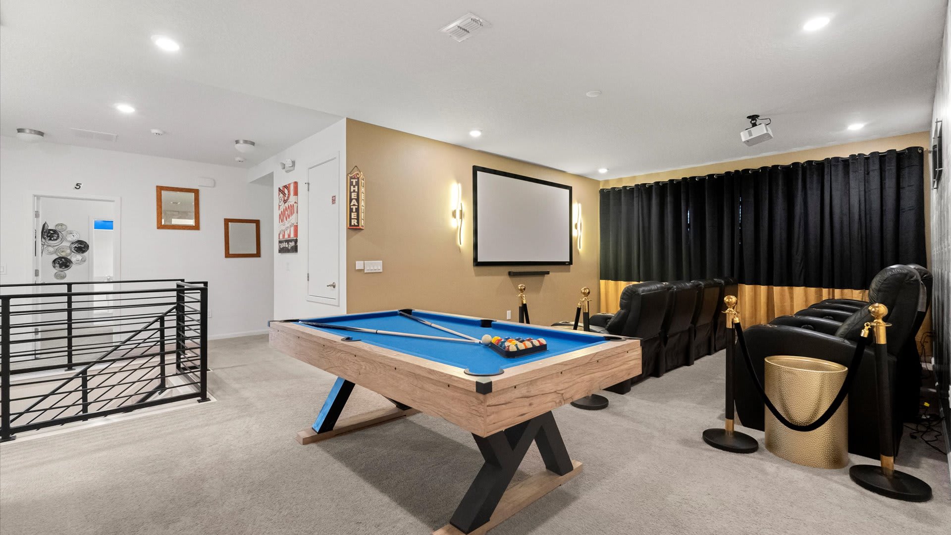 An expansive entertainment space combines a home theater with a pool table, featuring black reclining seats, a large projection screen, and a light wood pool table with blue felt, all visible from a carpeted landing with a modern metal railing.