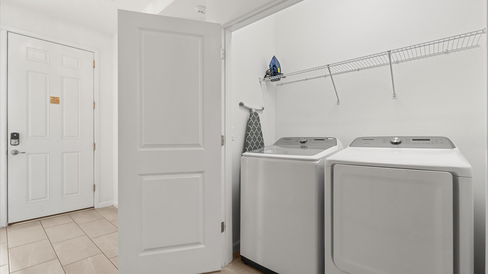 Bright laundry room featuring a white Samsung washer and dryer, a wire shelf, and an ironing board, with a white door leading to another area of the house.