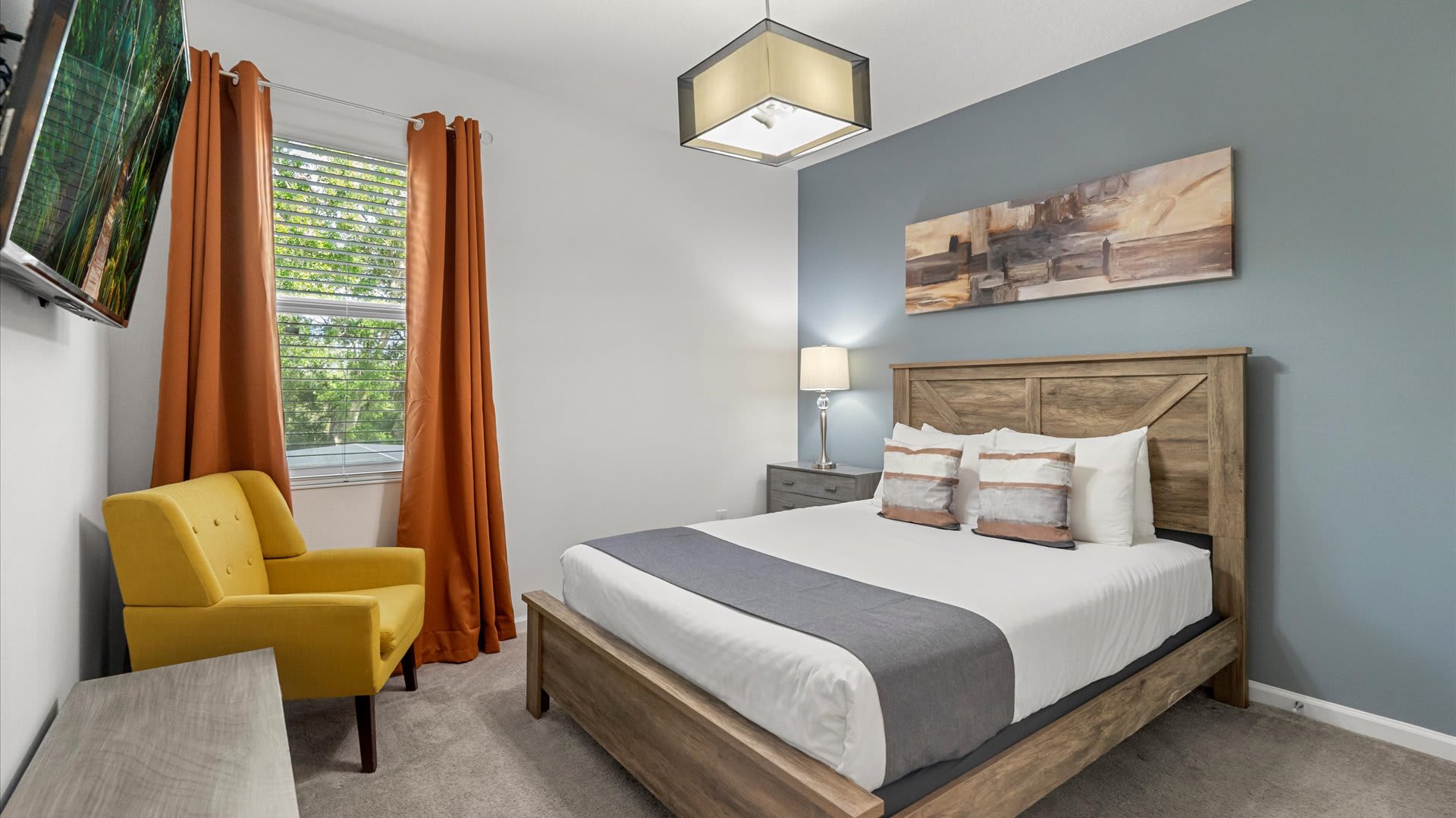 Cozy bedroom featuring a wooden queen-sized bed with white and gray bedding, a nightstand with a lamp, and a bright yellow armchair. An orange curtain frames the window, and a blue accent wall displays abstract art.