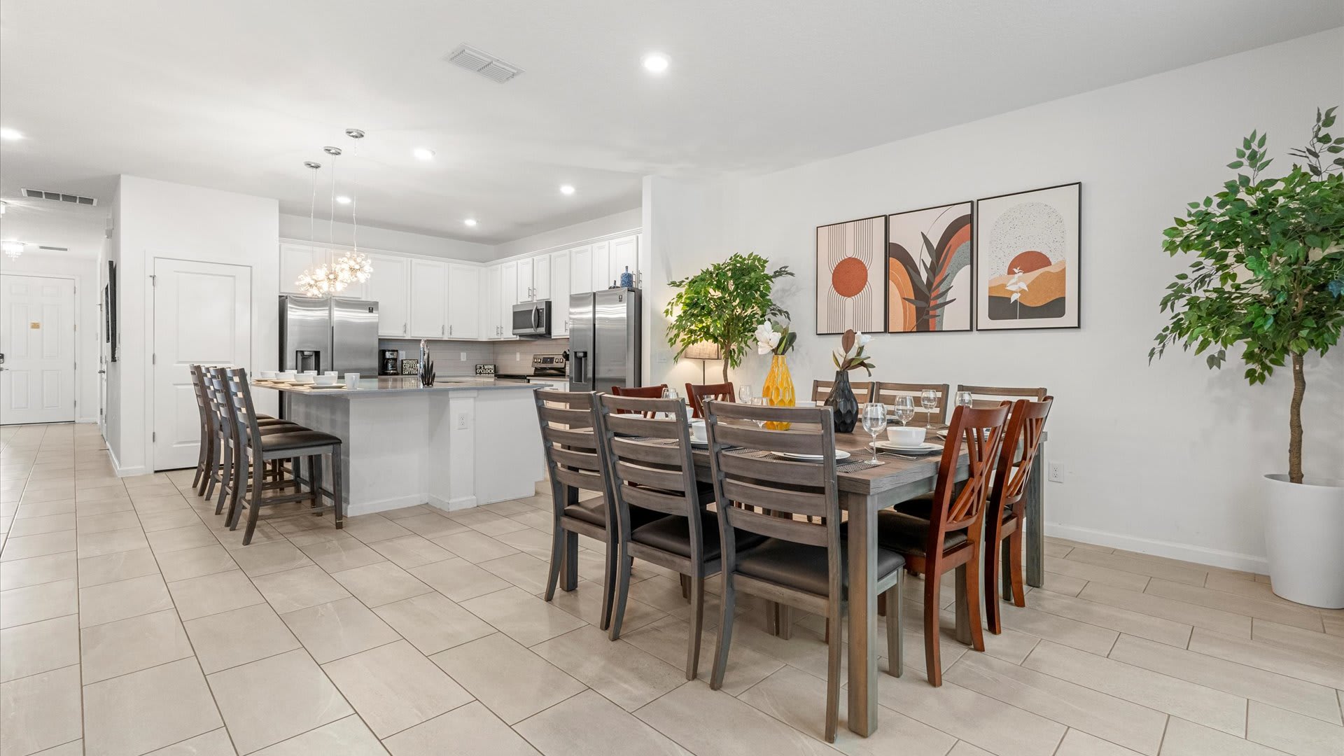 An open-concept dining area with a large wooden table and chairs, set against a white wall with three abstract art pieces, flowing into a modern kitchen with white cabinets and stainless steel appliances.