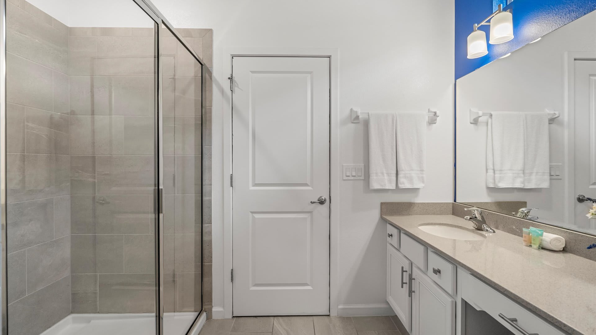 Modern bathroom with a walk-in glass shower featuring light gray tiled walls, a single vanity with a mirror, and white towels hanging on the wall.