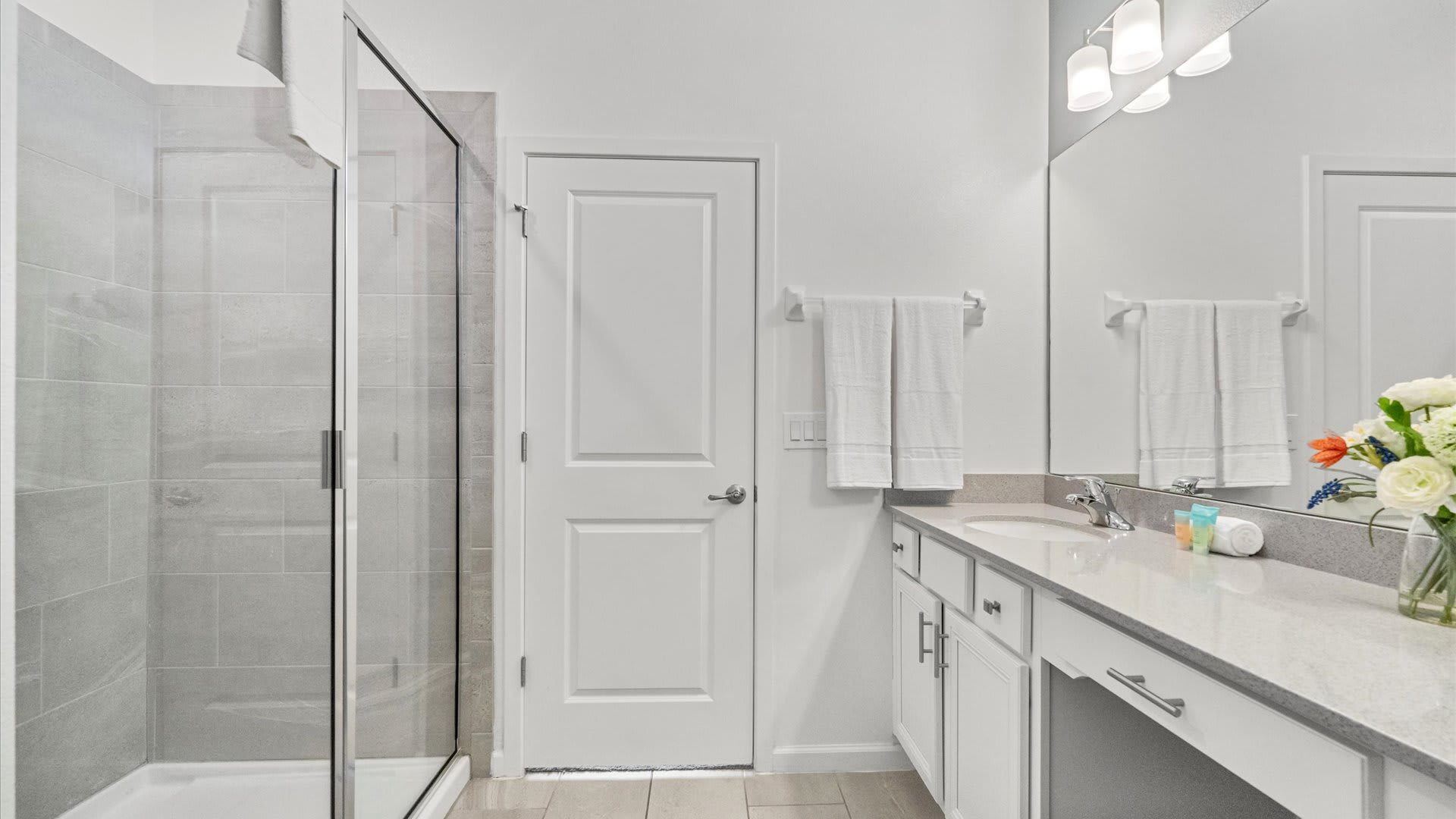 Bright bathroom with a glass-enclosed shower featuring gray tile, a long single vanity with a light gray countertop, and a large mirror. White towels hang on the wall next to a white door.