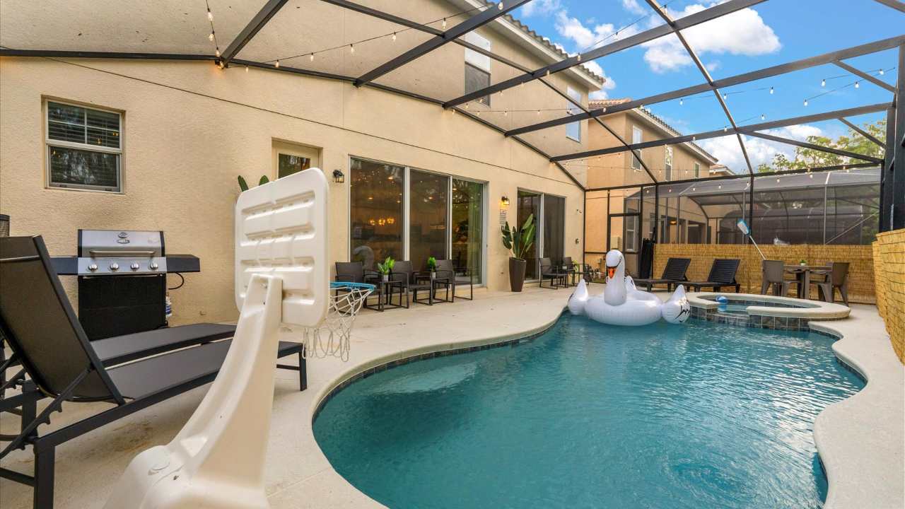 An inviting outdoor pool area enclosed by a screen cage, featuring a curved swimming pool with a large inflatable swan. There's a basketball hoop, lounge chairs, a BBQ grill, and an outdoor dining set on the patio.