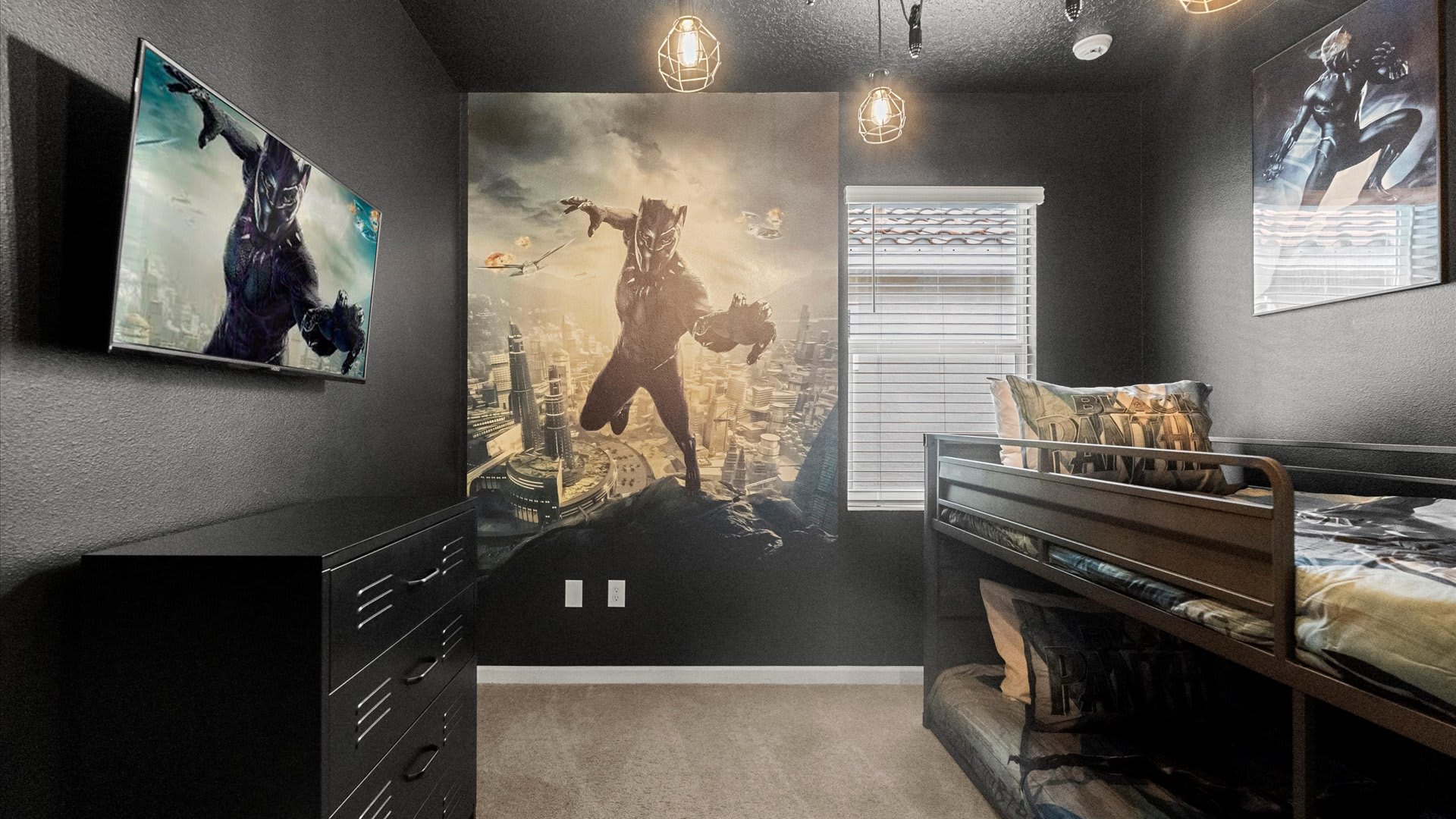 Themed kids' bedroom with a large Black Panther mural, a wall-mounted TV, a black dresser, and a unique black metal bunk bed designed to resemble a shipping container.
