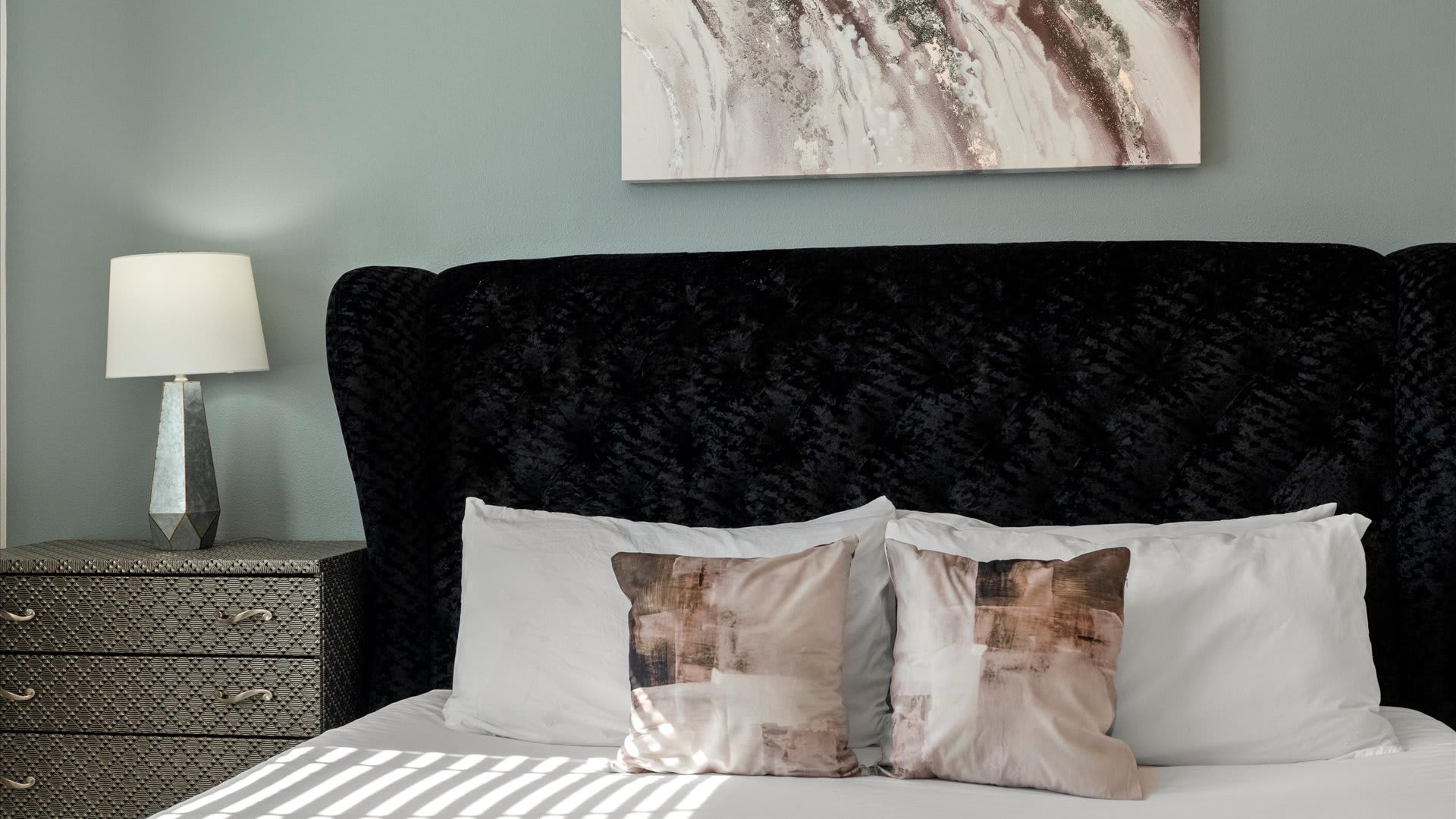 A cozy bedroom featuring a luxurious black velvet tufted headboard on a bed with white and patterned pillows, complemented by a dark patterned nightstand and a modern table lamp.
