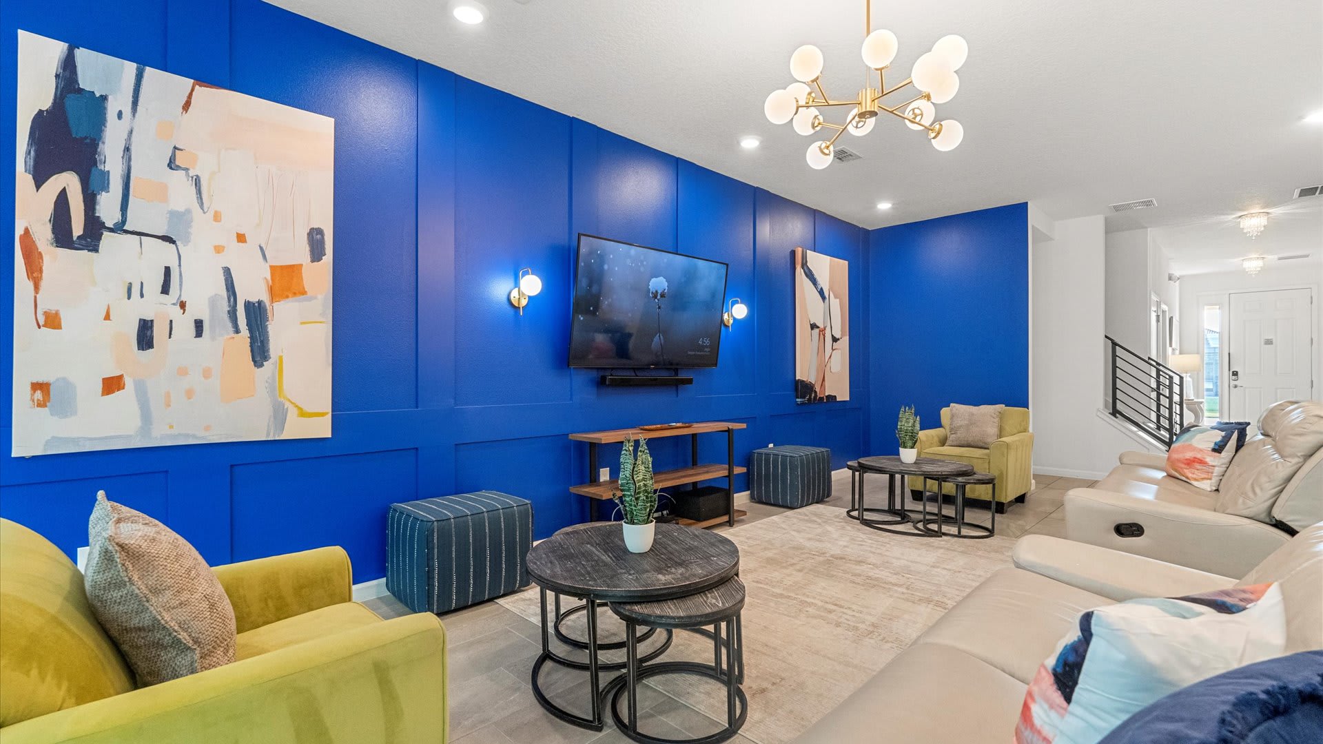 The bright living room is anchored by a bold blue paneled accent wall with a wall-mounted TV and abstract art, complemented by a light-colored sectional sofa, yellow armchairs, and modern nesting coffee tables on a light rug, with a contemporary chandelier overhead.