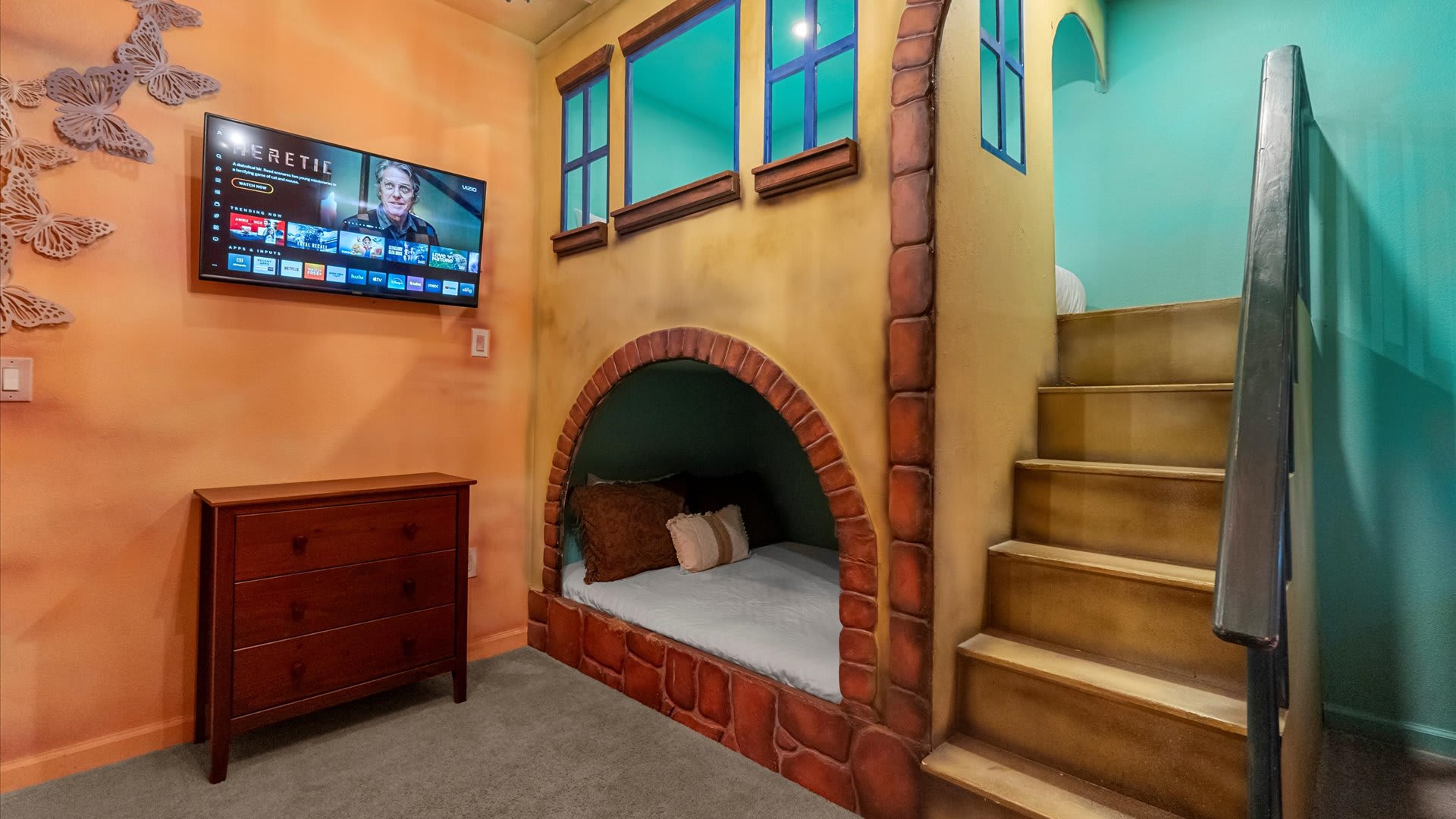Close-up view of the themed bedroom showing a built-in bed with an arched brick-like entrance, a wooden dresser, and a wall-mounted TV. Butterfly decals decorate the orange wall.
