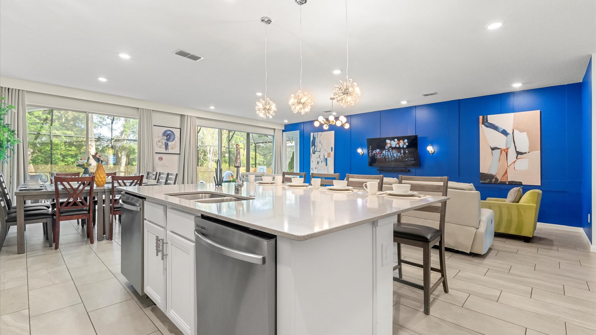 An expansive open-concept space connects the kitchen island with bar seating to a dining area with a large wooden table, leading into a vibrant living room with a blue accent wall and large sliding glass doors offering views to the outdoors.