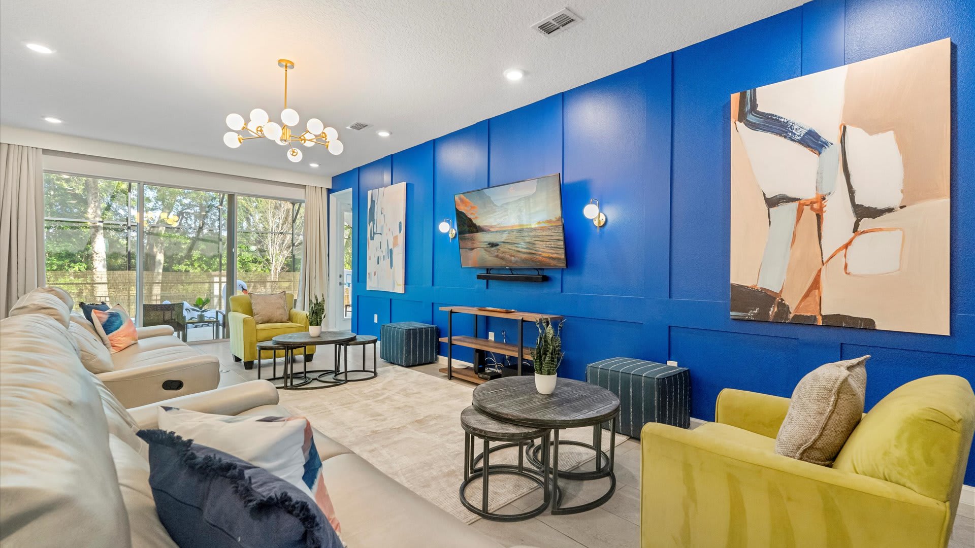 A modern living room features a striking royal blue accent wall with paneling, showcasing a wall-mounted TV flanked by sconces and large abstract artwork, with a light sofa and yellow armchairs arranged around nesting coffee tables.