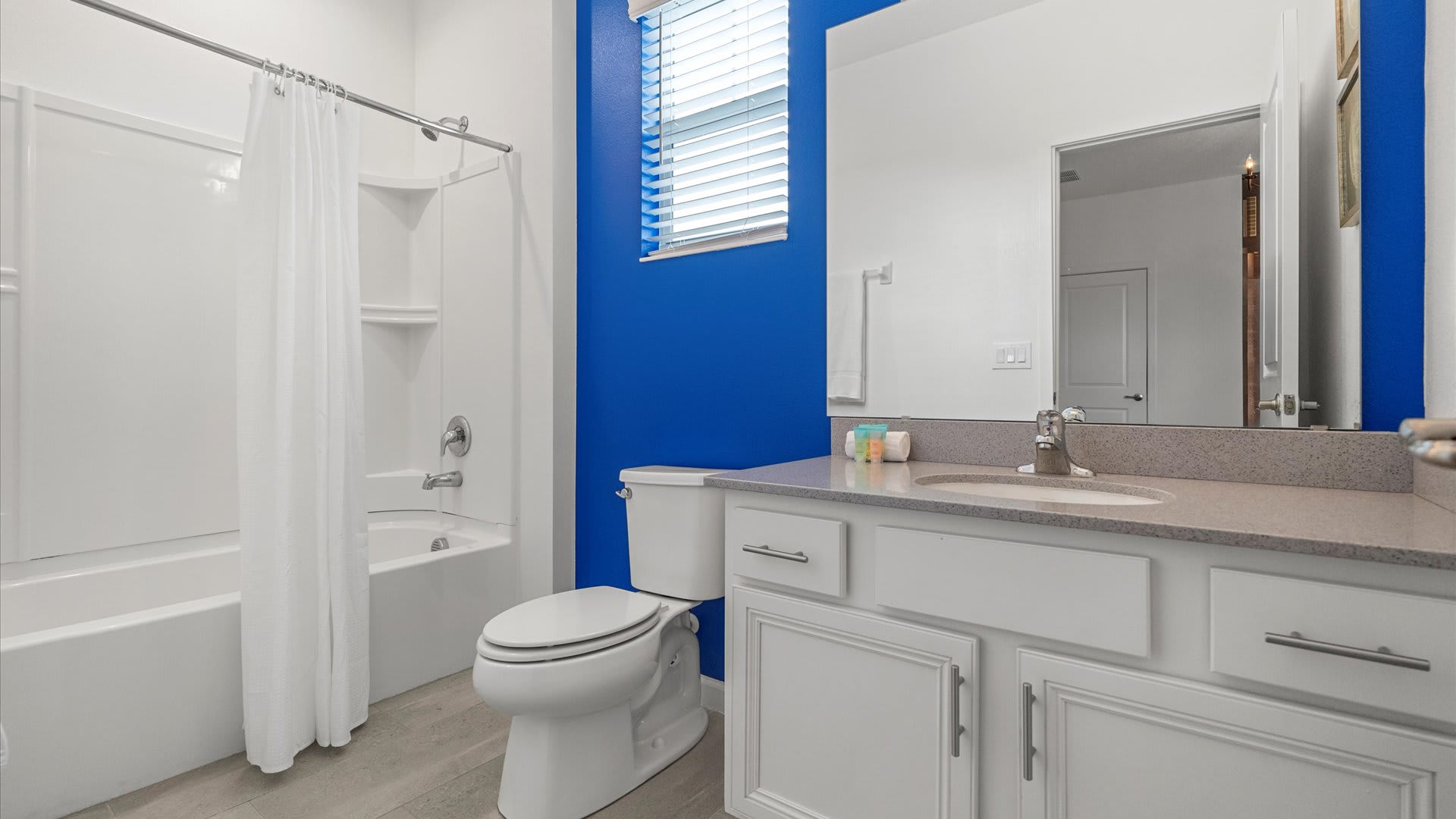 Clean white bathroom with a single vanity, a toilet, and a shower/tub combo, highlighted by a bold blue accent wall behind the toilet and vanity area.
