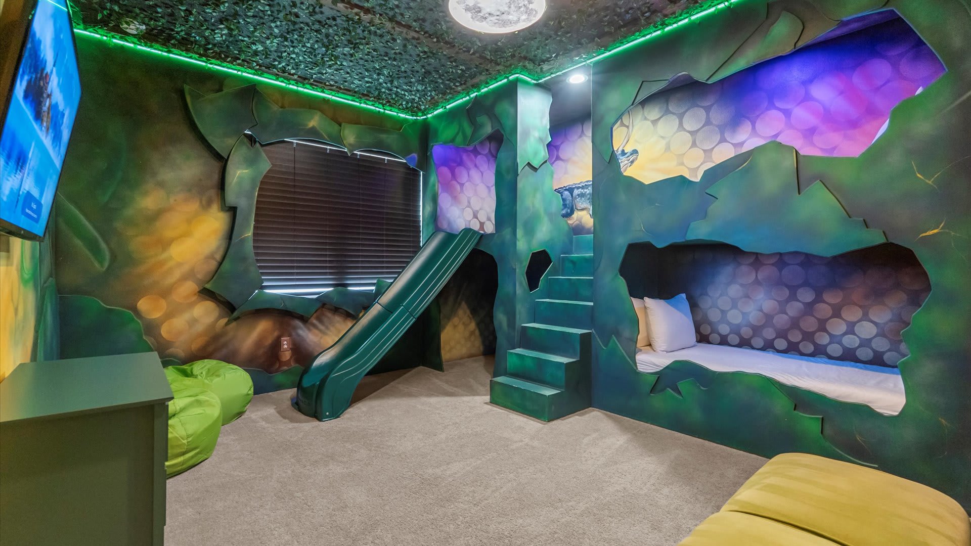 Whimsical themed bedroom designed like a jungle cave with green walls, built-in bunk beds, and a green slide. The ceiling is covered in leafy decor, and a wall-mounted TV is visible.