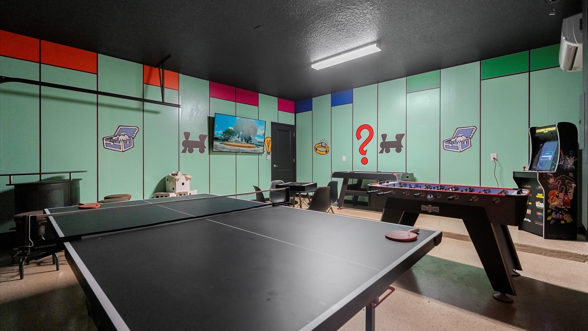 A vibrant game room with light green walls painted with Monopoly board game elements and a dark ceiling. It features a ping-pong table in the foreground, a foosball table, a pool table, and an arcade machine.