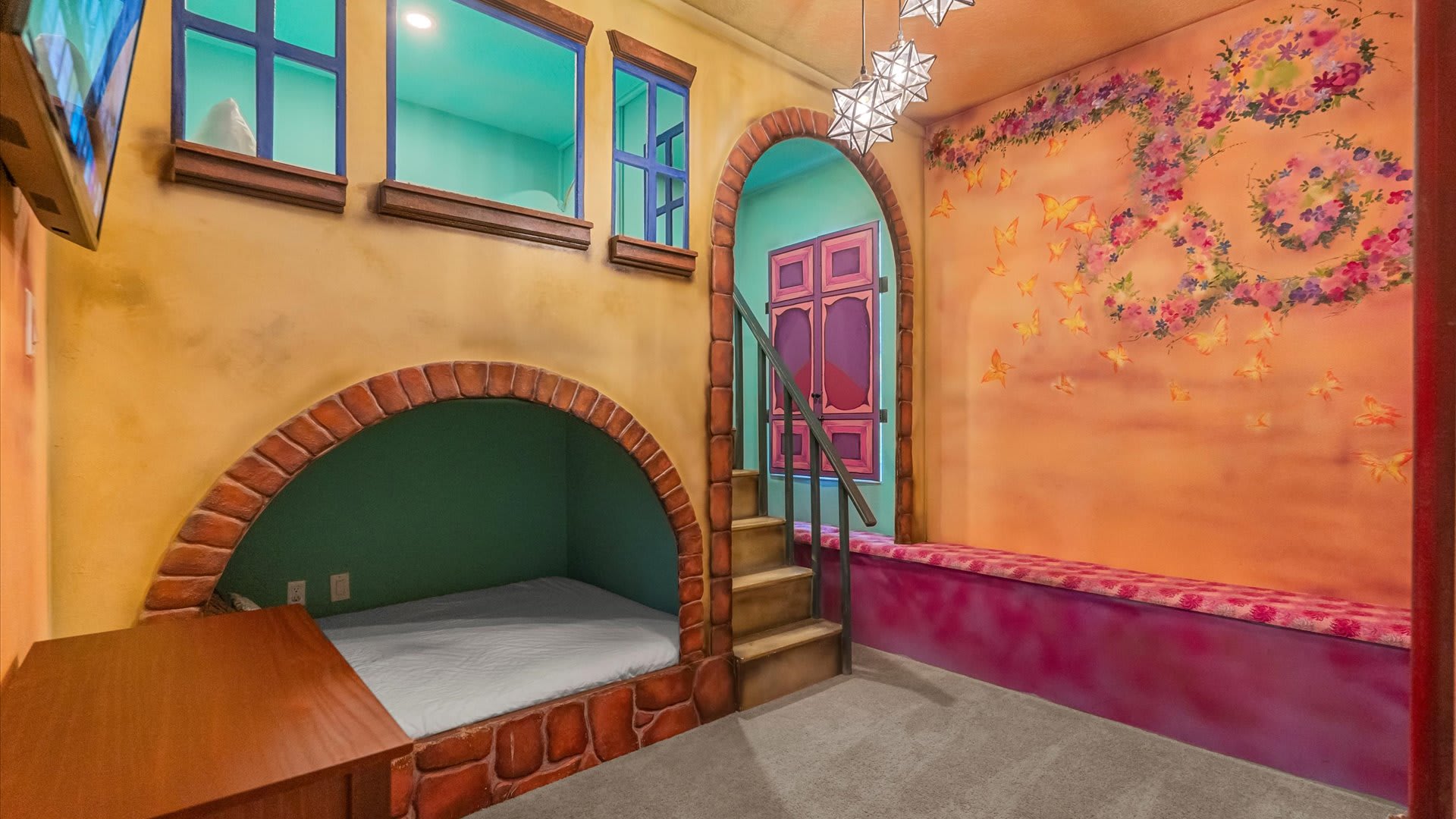 Vibrant themed bedroom with built-in beds resembling a colorful house facade, complete with arched openings and windows. Steps lead to an upper sleeping area, and the walls are adorned with floral and butterfly murals.
