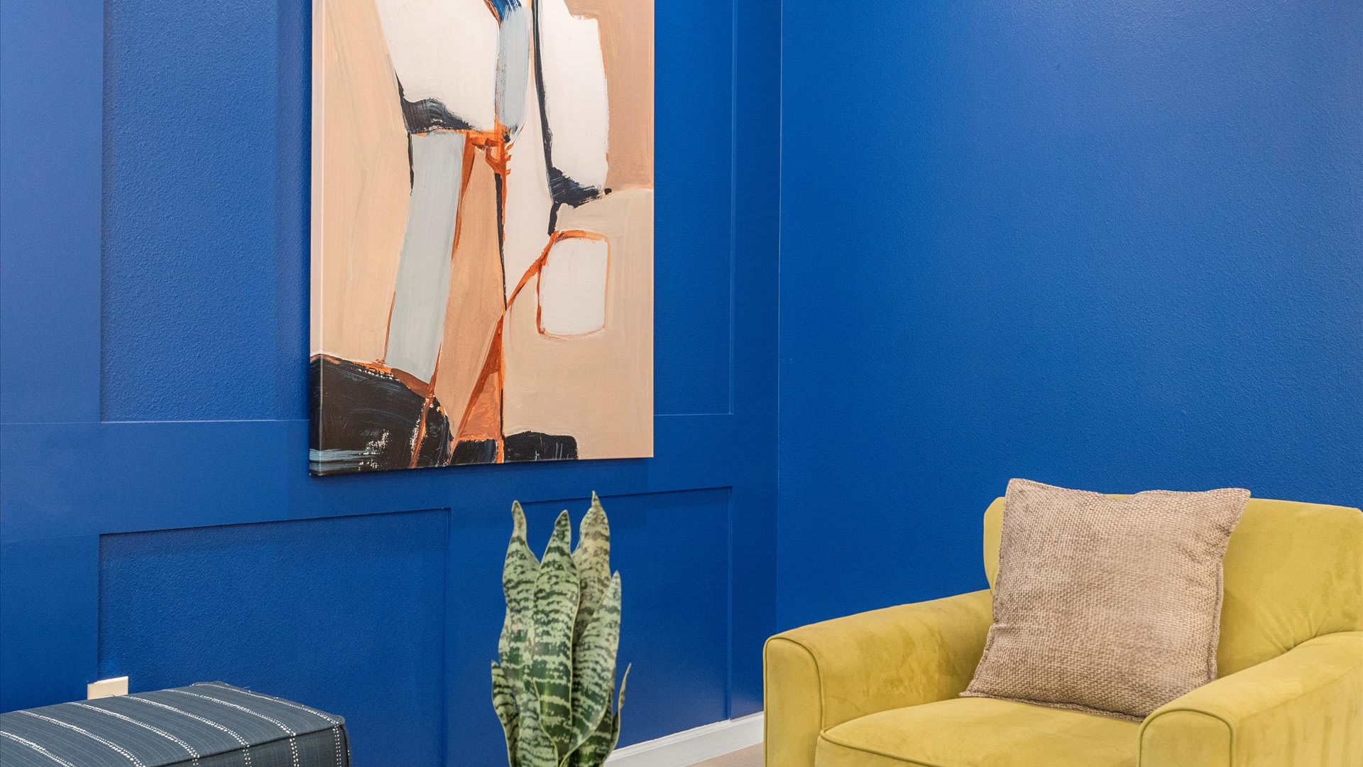 A close-up view in the living room highlights a comfortable yellow armchair positioned against the vibrant blue paneled accent wall, beneath a large abstract painting, with a small snake plant on a side table.