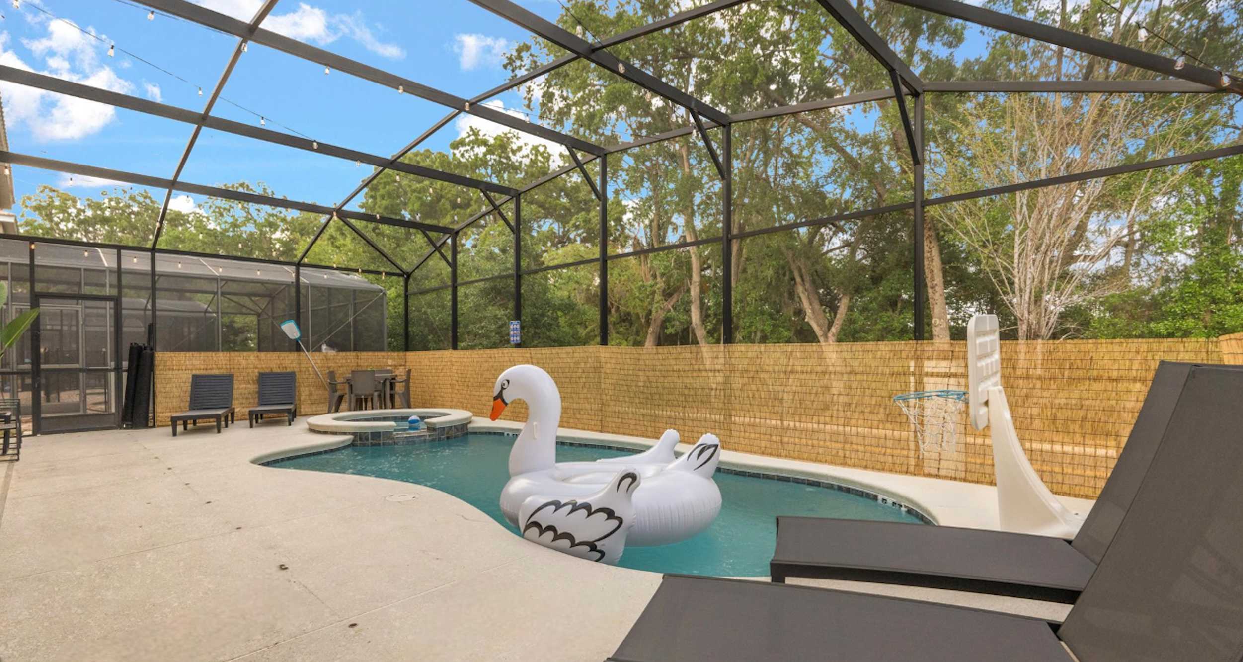 A wide view of the screened-in private pool area, featuring a large swimming pool with an inflatable swan and a hot tub. The area is surrounded by a low brick wall and lush trees, with string lights overhead.