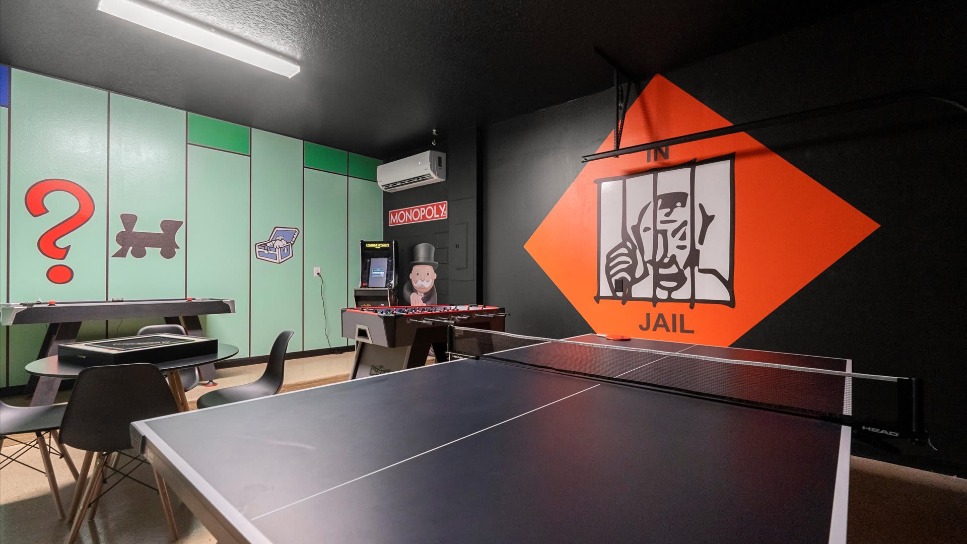 A vibrant game room with a Monopoly theme, featuring a large 'In Jail' mural on an orange and black wall. A pool table, foosball table, and arcade machine are visible, along with a round table and chairs.
