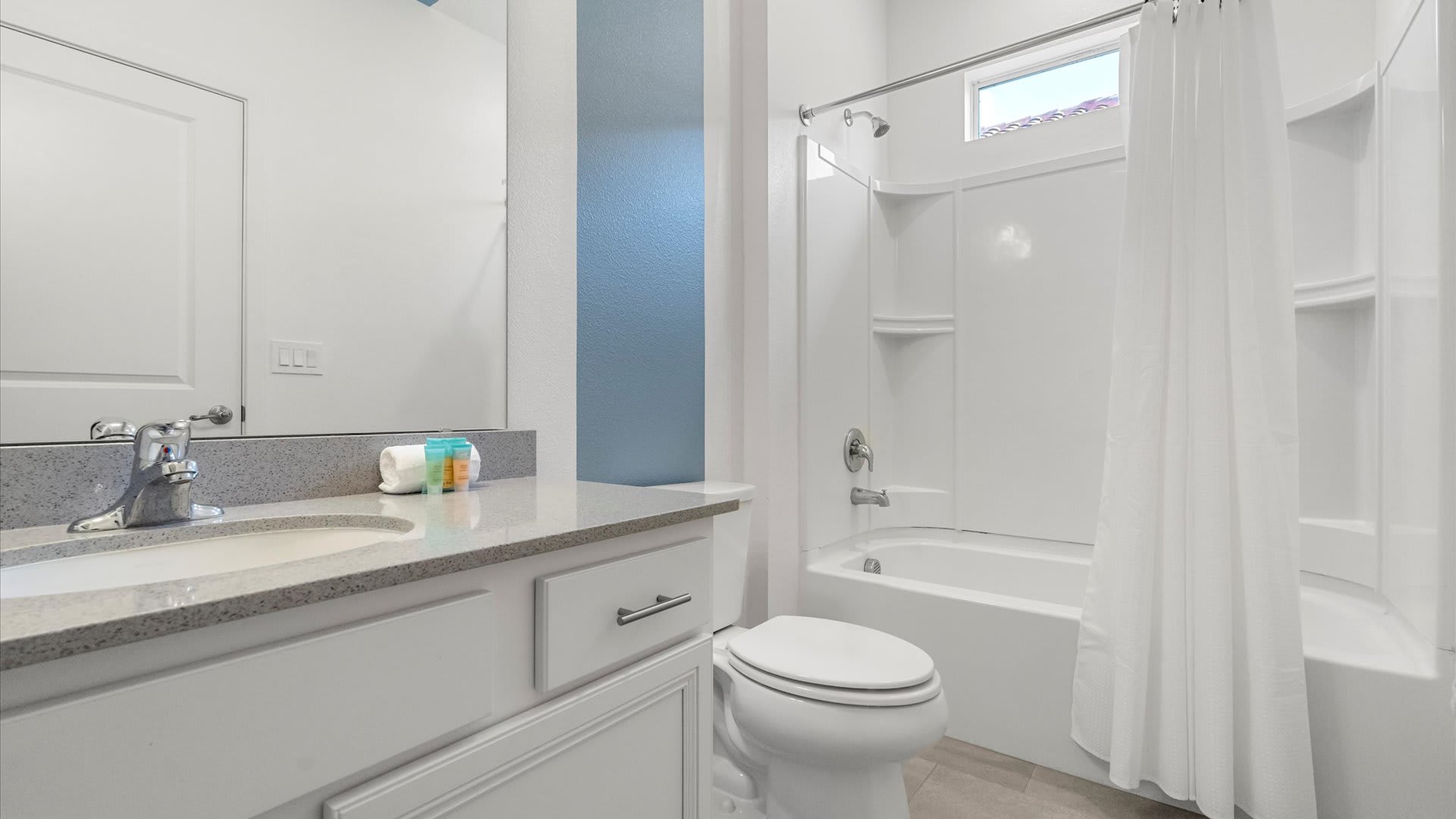 Modern white bathroom featuring a single vanity with a light gray countertop, a large mirror, a toilet, and a shower/tub combo with a white curtain. A vibrant blue accent wall adds a pop of color.