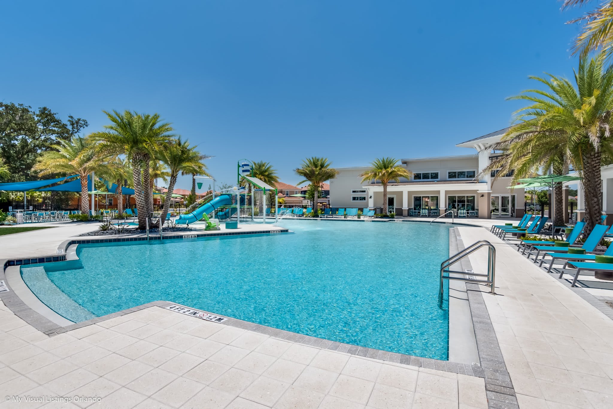 An expansive communal swimming pool with a wide paved deck, surrounded by palm trees and lounge chairs. A kids' splash pad with a blue slide is visible on the left, with a modern clubhouse building in the background under a clear blue sky.