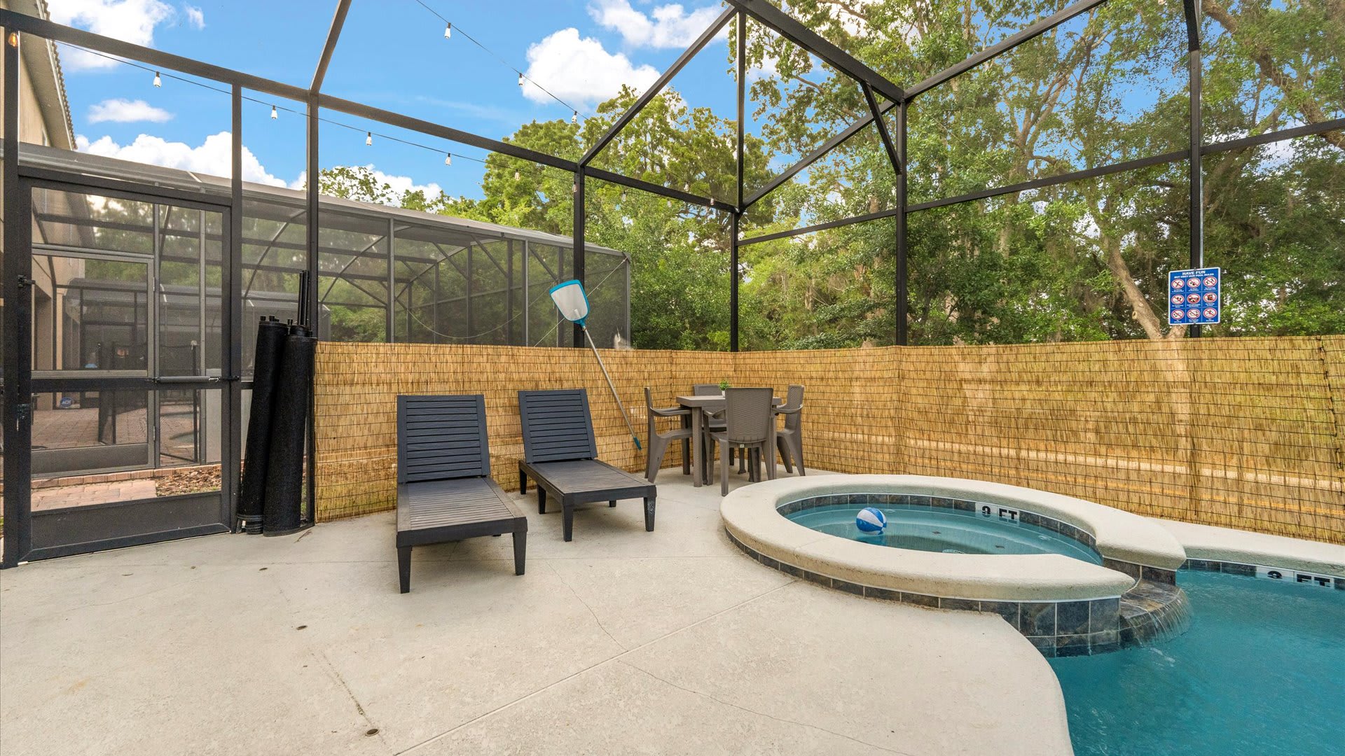 A closer view of the screened-in pool area, highlighting the integrated hot tub and a small outdoor dining set. Two dark lounge chairs are visible, and a bamboo-style privacy screen lines the back wall.