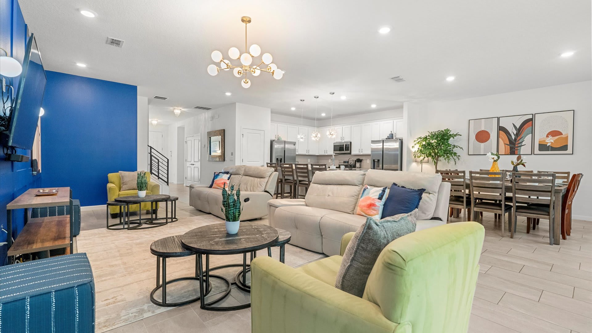 A bright, open-concept living space with light tile flooring, a royal blue accent wall, and two light-colored reclining sofas. A modern gold chandelier hangs above, and a dining area with a wooden table and chairs is visible in the background, next to a white kitchen.