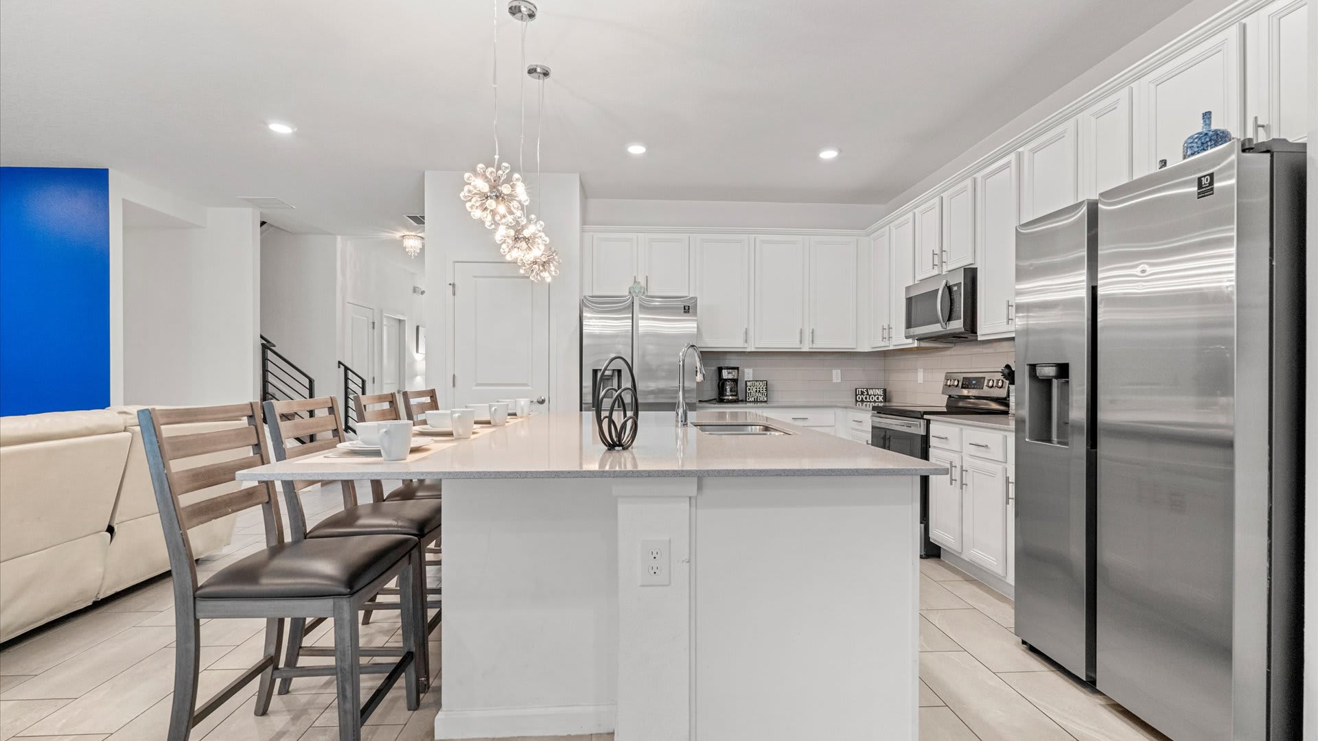 The open-concept kitchen features a large island with a sink and bar seating for four, illuminated by modern pendant lights, surrounded by white shaker cabinets and stainless steel appliances, seamlessly connecting to the living and dining areas.