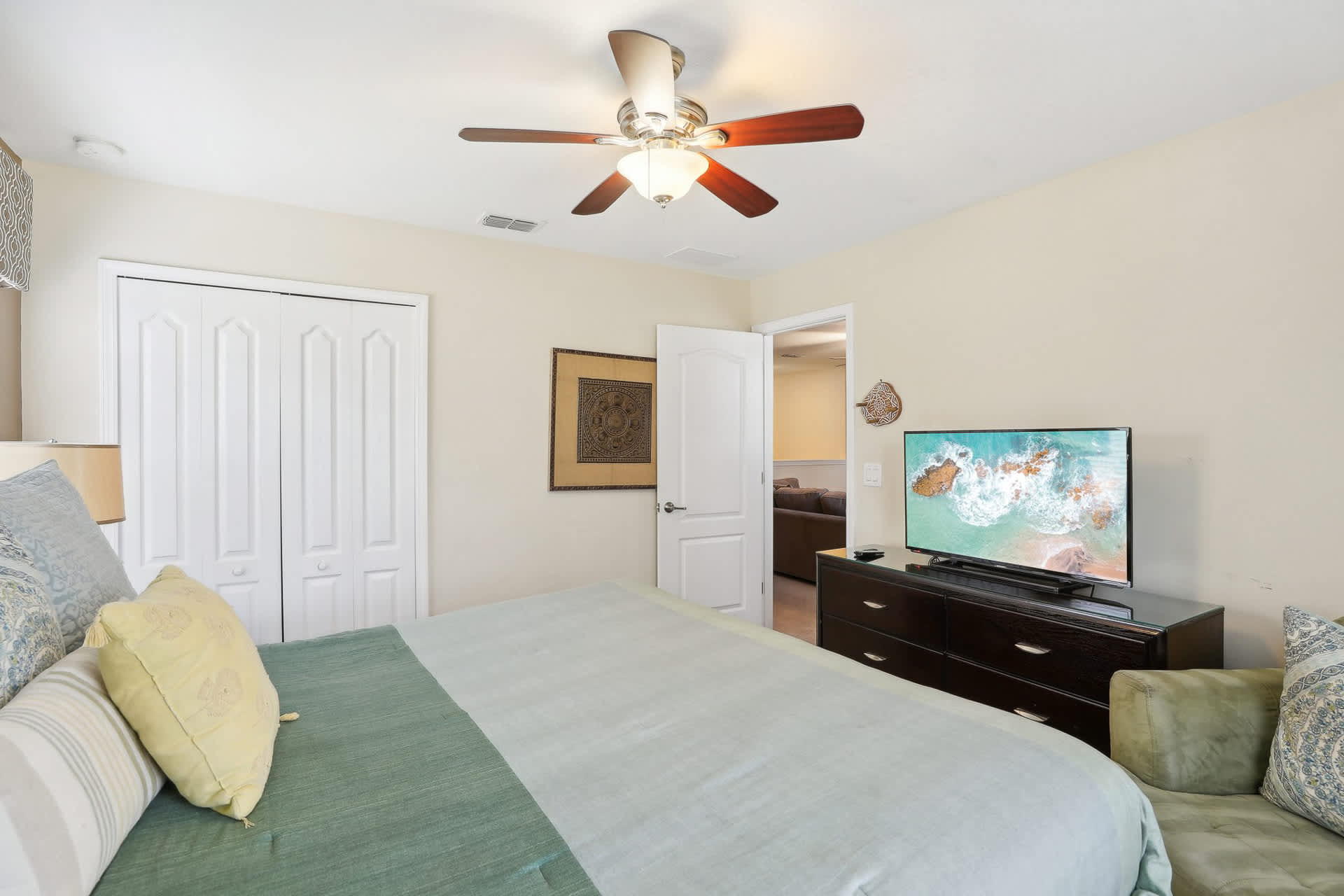 View of the bedroom with a large flat-screen TV on a dark wood dresser, a king-sized bed with green bedding, and an open door leading to another room.