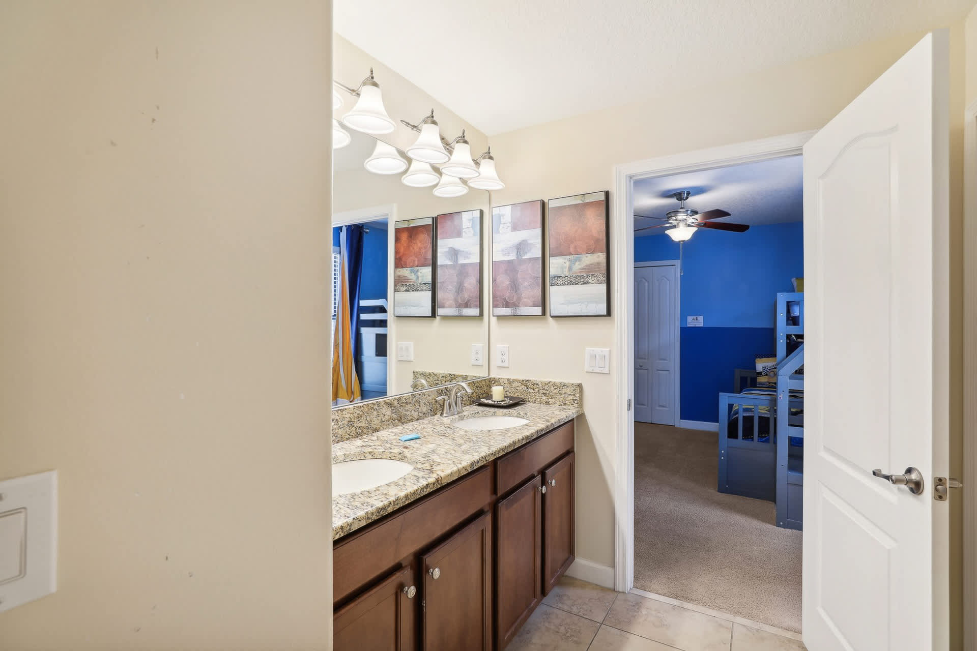 A bright bathroom vanity area features a double sink with a granite countertop, dark wood cabinets, and a large mirror reflecting a bedroom with blue walls and a ceiling fan.