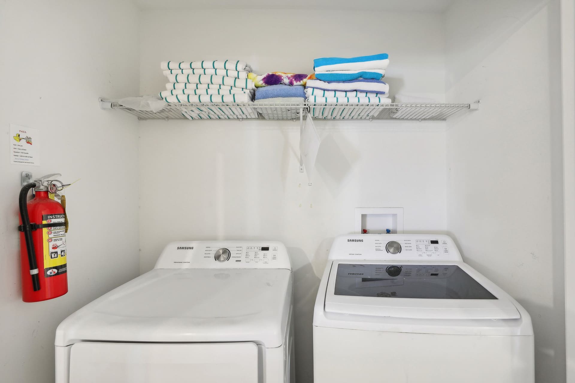 A compact laundry room features a white Samsung washer and dryer, a wire shelf stocked with neatly folded towels, and a red fire extinguisher mounted on the wall.