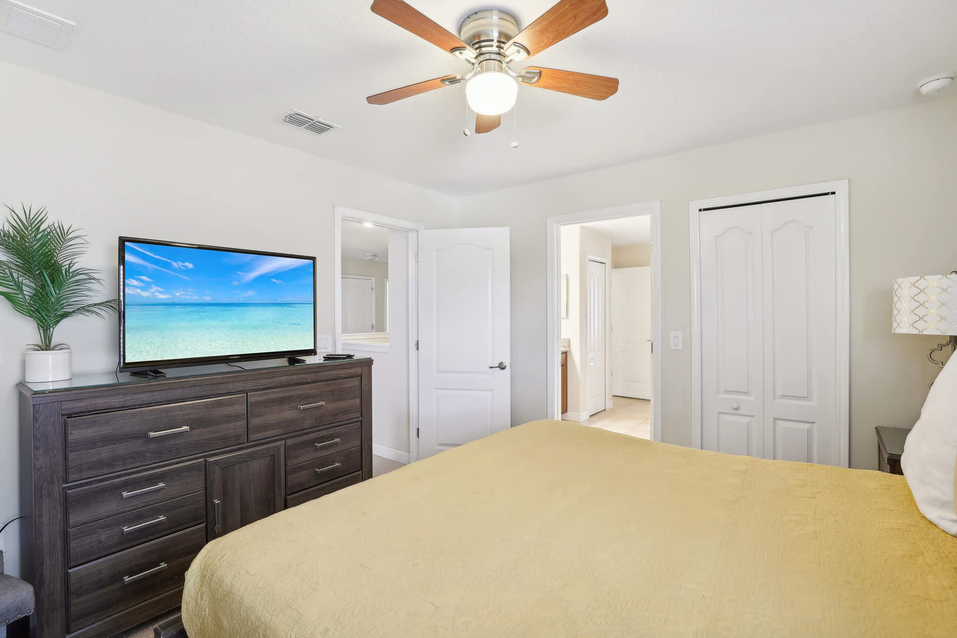 Spacious bedroom featuring a dark wooden dresser with a large flat-screen TV displaying a beach scene, a light yellow bedspread, and a ceiling fan with wooden blades.